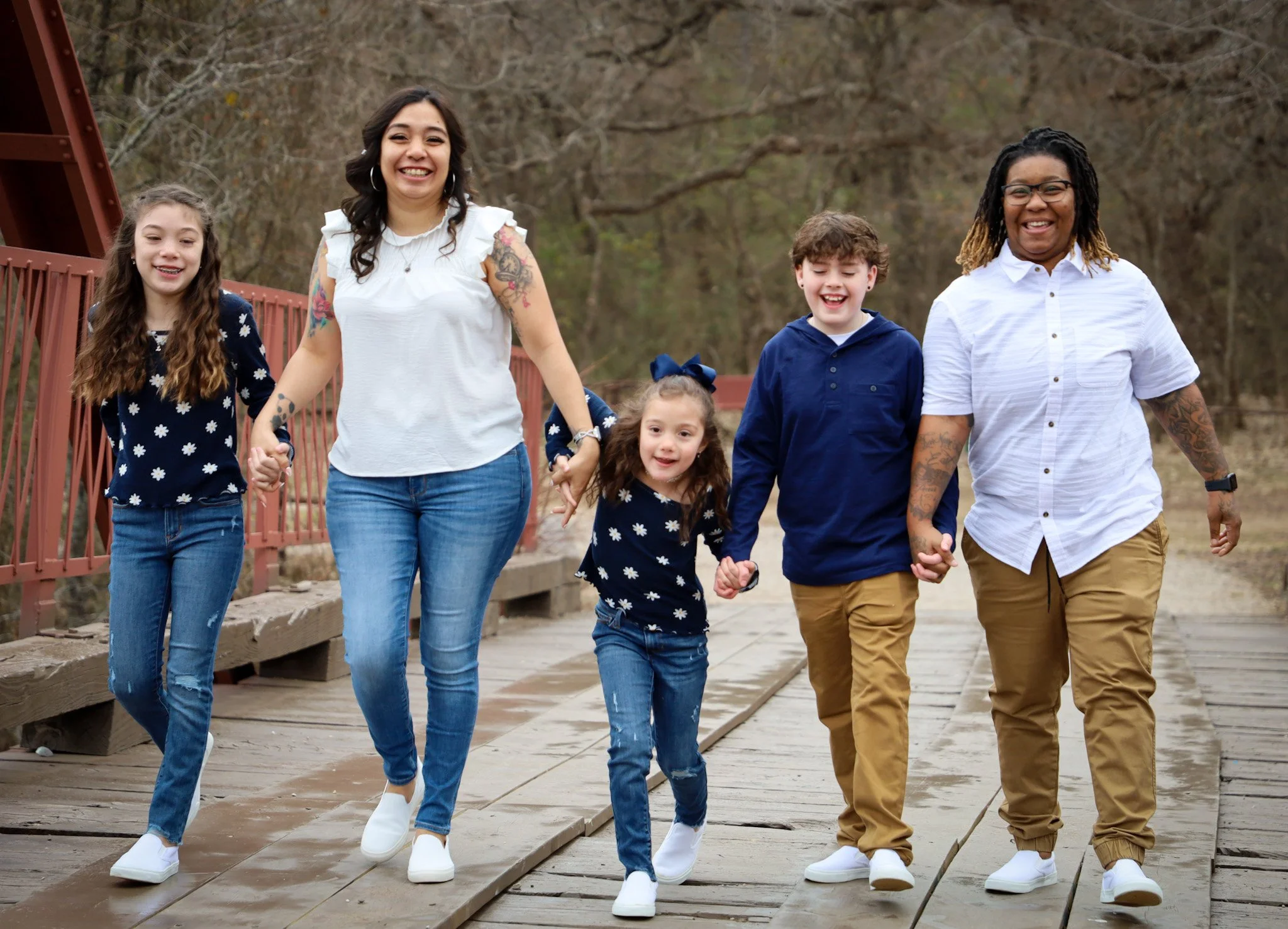 Group of six people, three children and three adults, walking hand in hand outdoors on a wooden pathway in a park during daytime with trees in the background, smiling and enjoying each other's company.