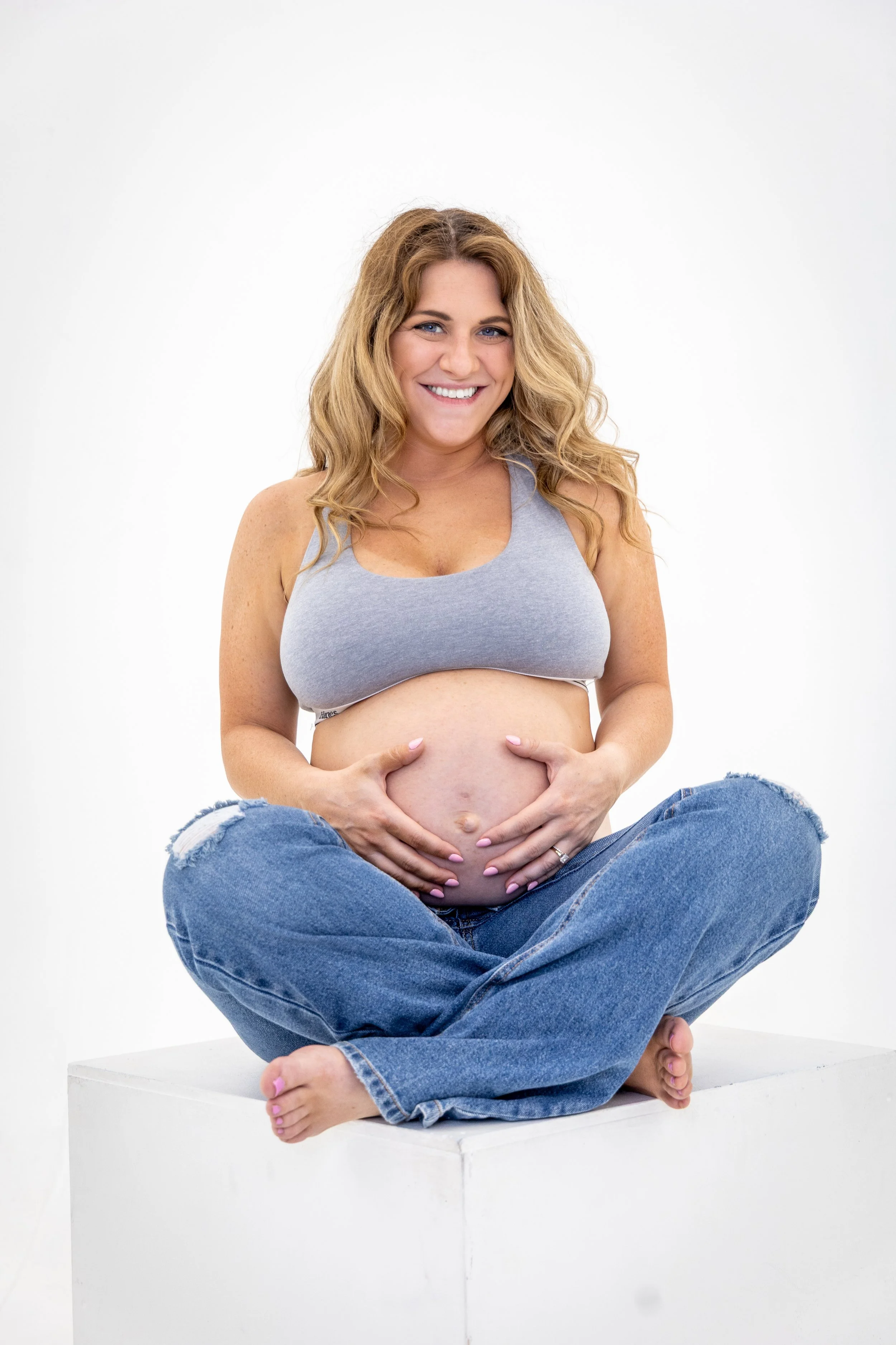 A pregnant woman with long wavy blonde hair sits cross-legged on a white platform against a plain white background. She is wearing a gray sports bra and ripped blue jeans, smiling, with both hands gently cradling her pregnant belly.