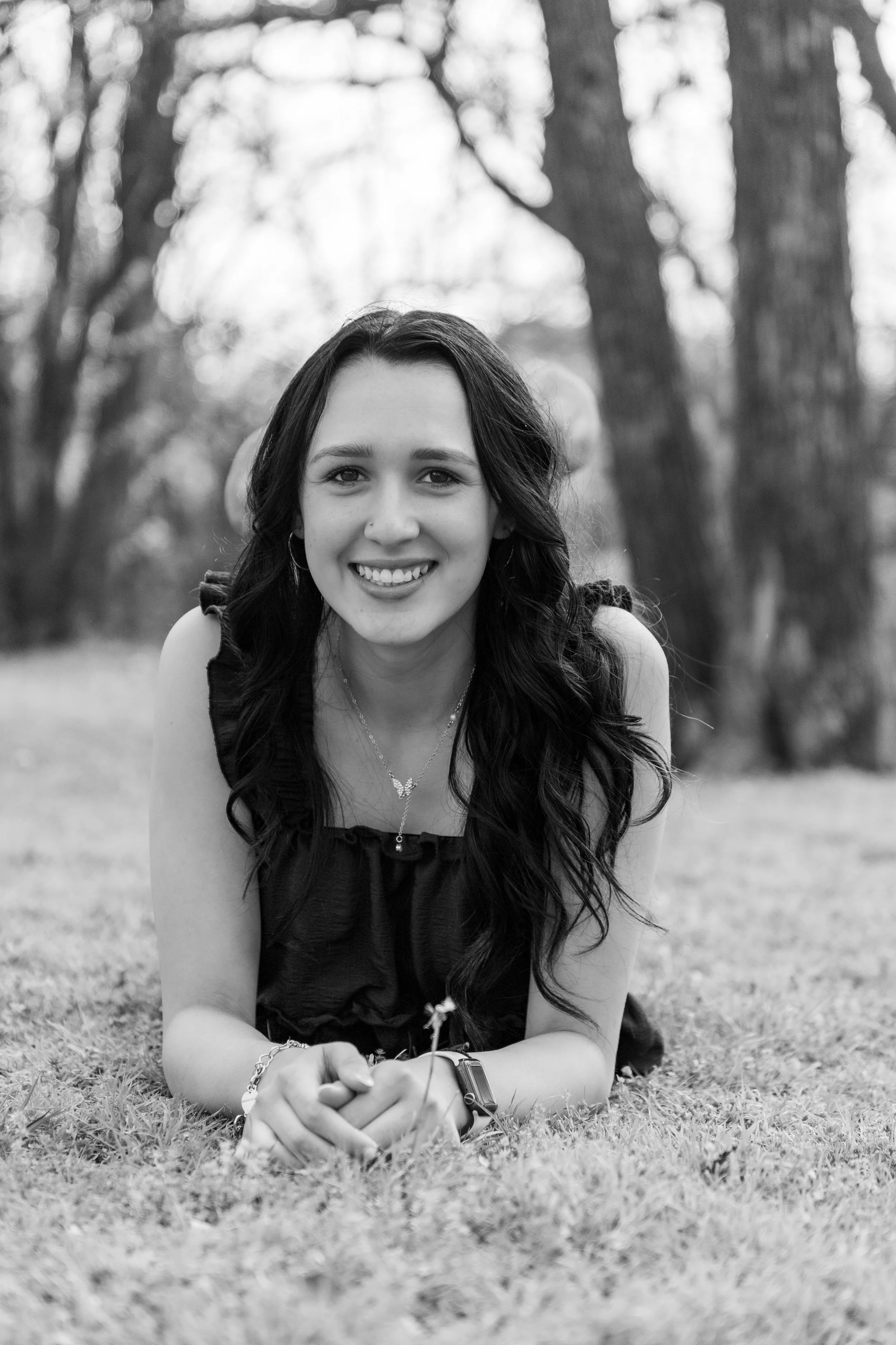 A young woman with long dark hair lying on grass in a park, smiling at the camera in black and white.