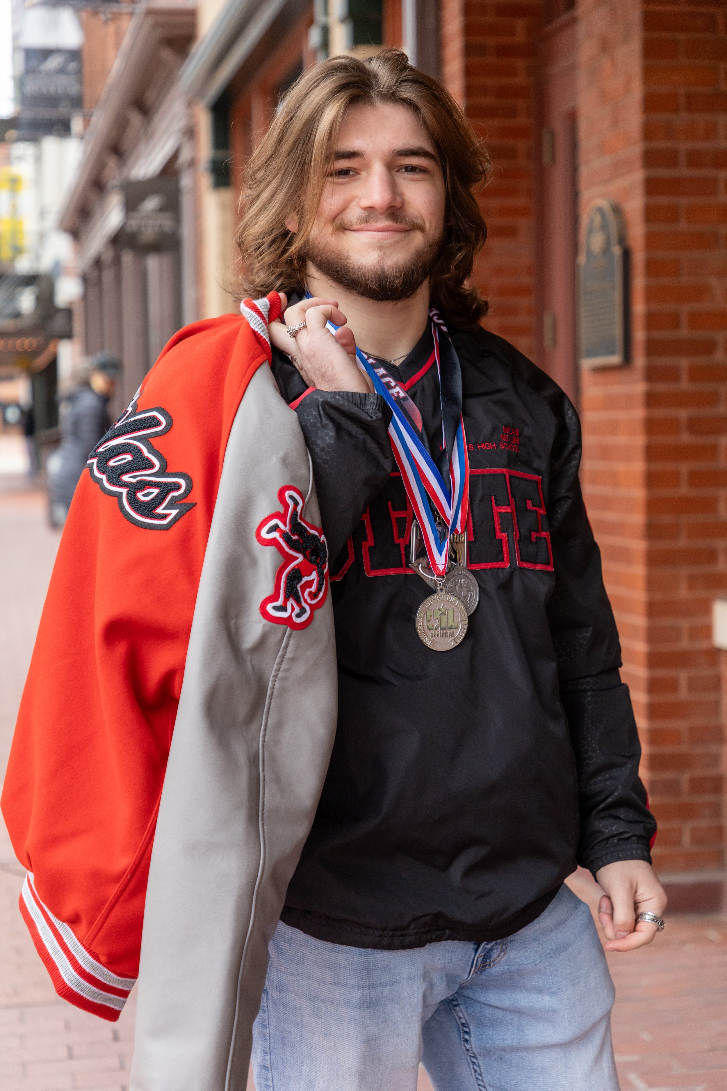 A young man with long brown hair and a beard, smiling, wearing a black jacket with red and gray accents, and losing a varsity jacket over his shoulder. He has medals around his neck and is standing on a city street with brick buildings in the backgro