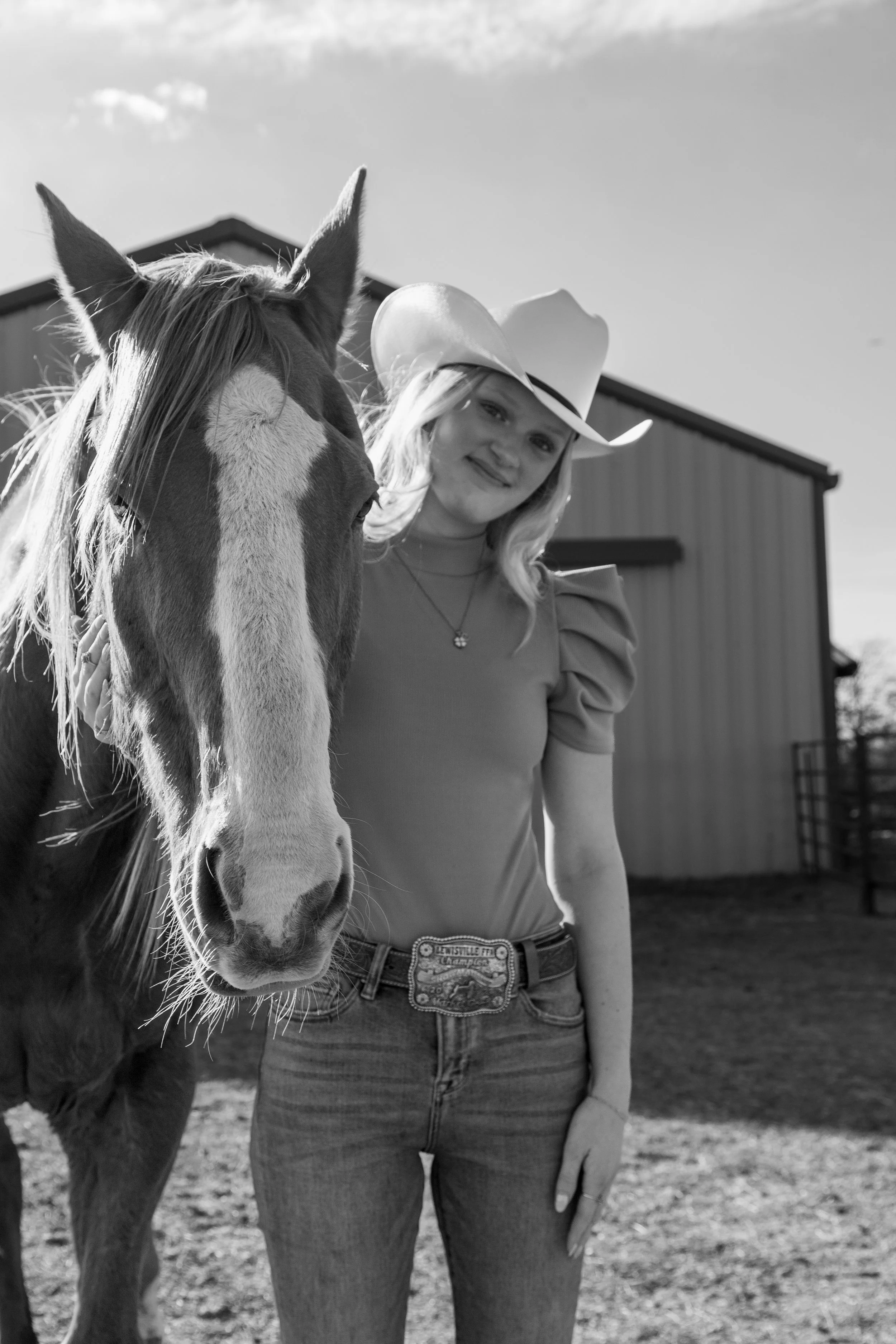 A woman wearing a cowboy hat and jeans stands next to a horse, outdoors near a barn.
