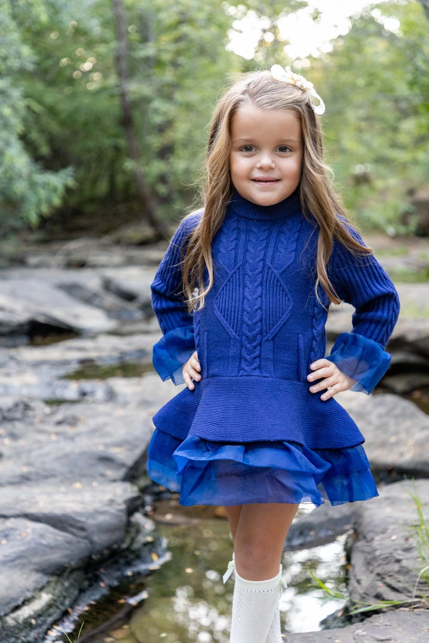 A young girl with long, wavy light brown hair and a white floral hair accessory, standing outdoors on rocks near a stream, wearing a royal blue knit sweater and a matching blue ruffled skirt, with white knee-high socks.