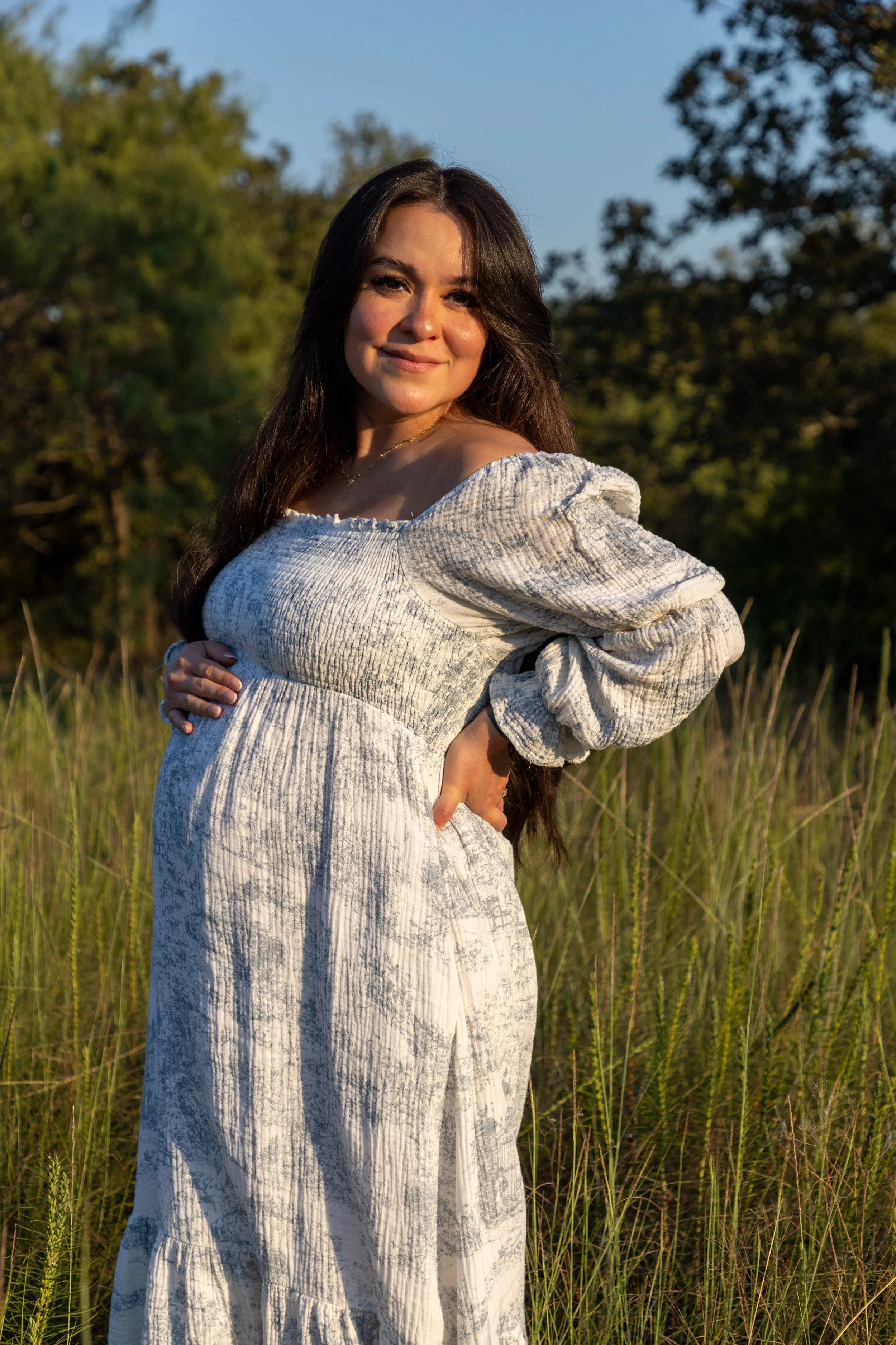 A pregnant woman with long dark hair standing in a grassy field during sunset, smiling and wearing a light-colored dress with puffy sleeves.