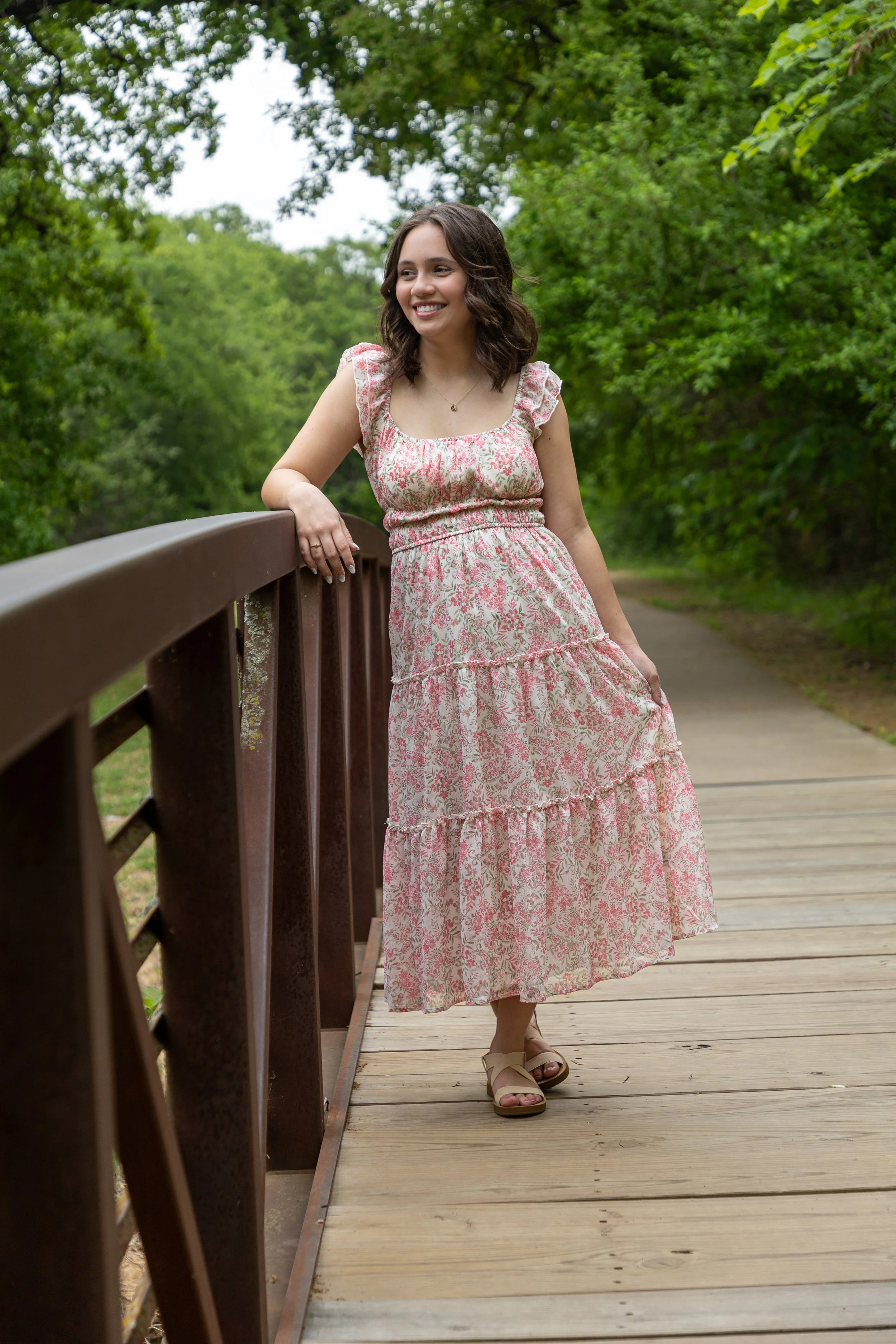 A young woman in a pink and white floral dress standing on a wooden bridge, surrounded by green trees in a park.