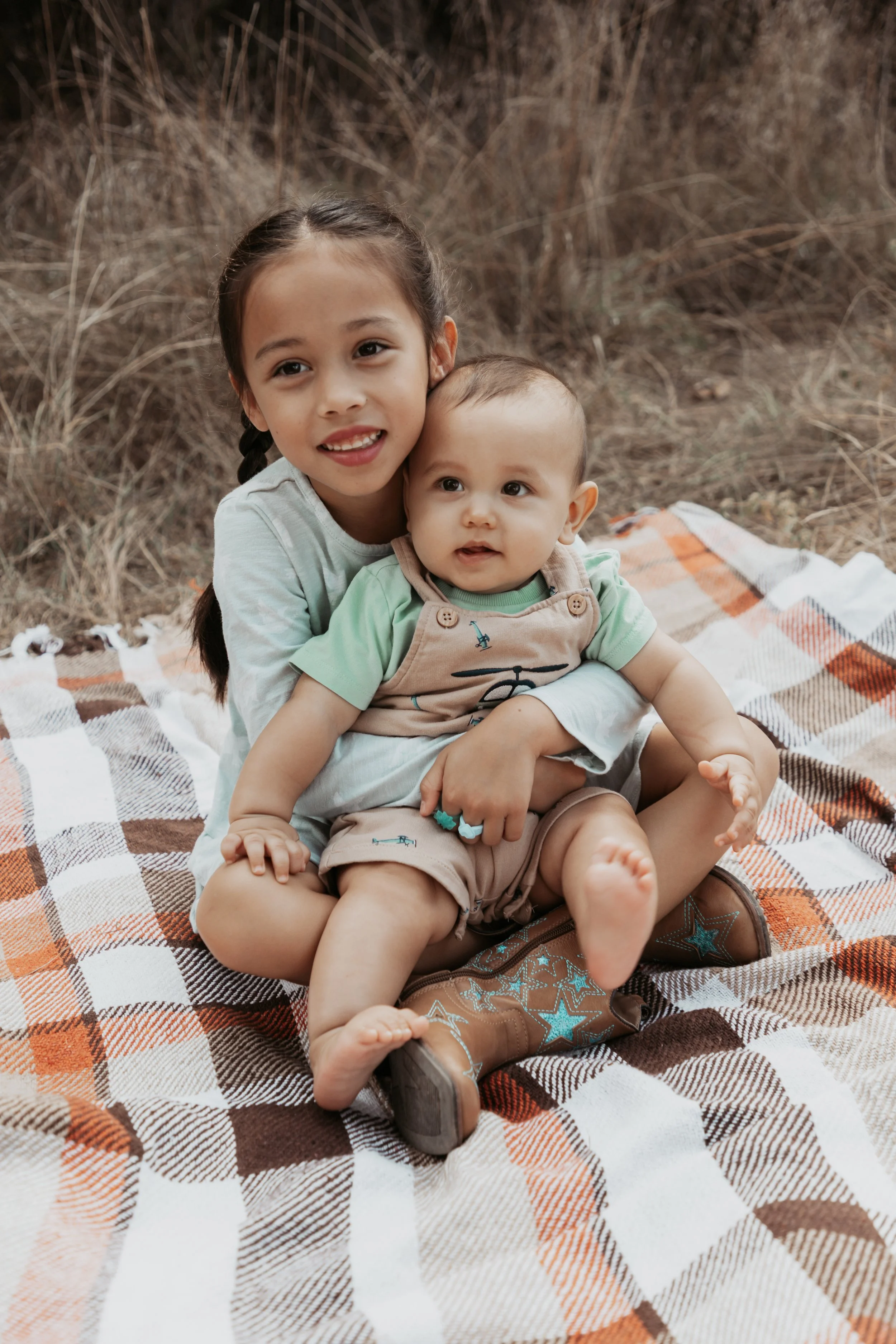 A young girl and a baby sitting on a checkered blanket outdoors, surrounded by dried grass during fall.