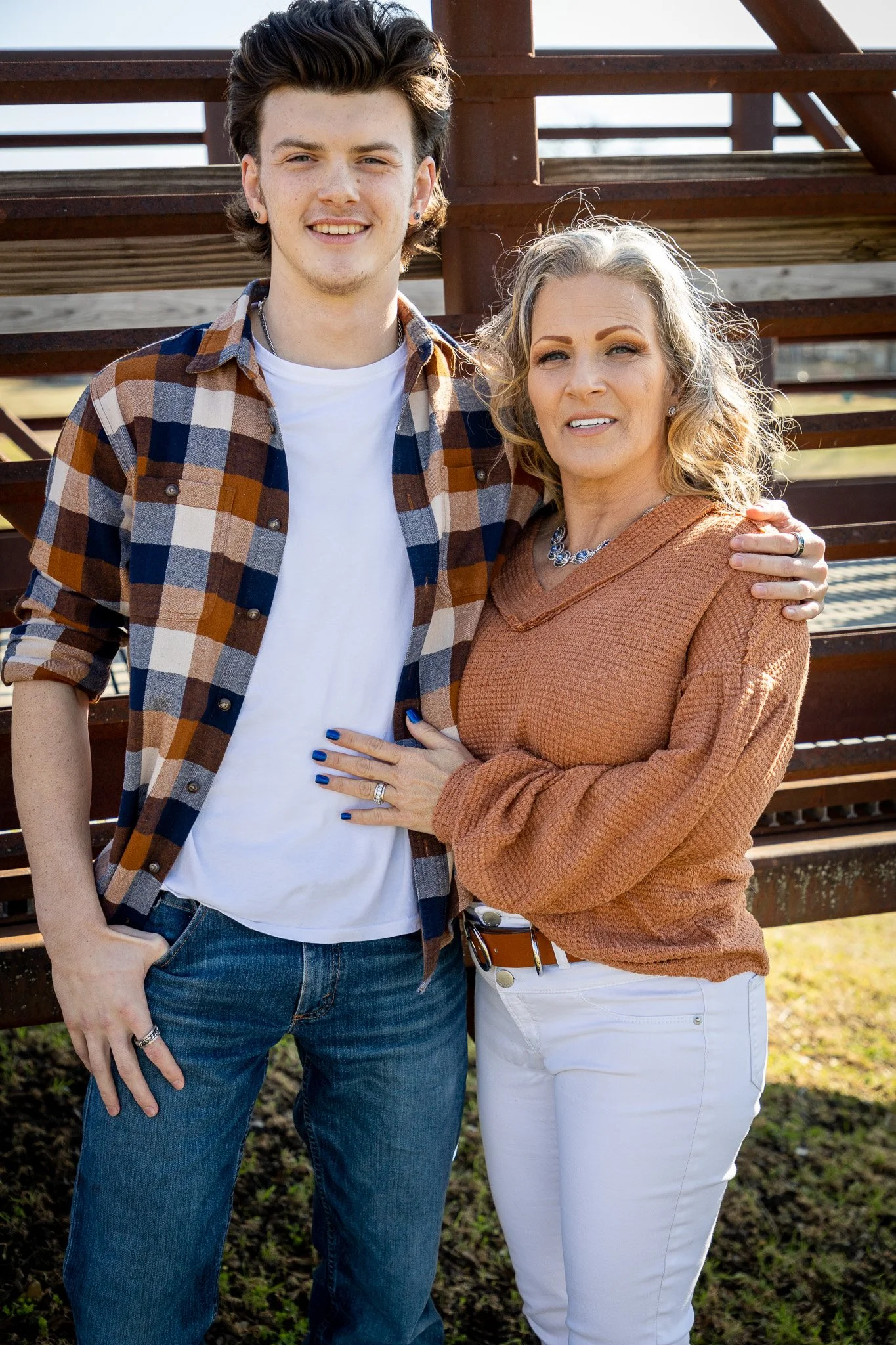 A young man and a woman stand outdoors in front of a wooden staircase, smiling and posing together. The man wears a plaid shirt, jeans, and a white t-shirt, while the woman wears a rust-colored sweater and white pants. The woman has her hand on the m