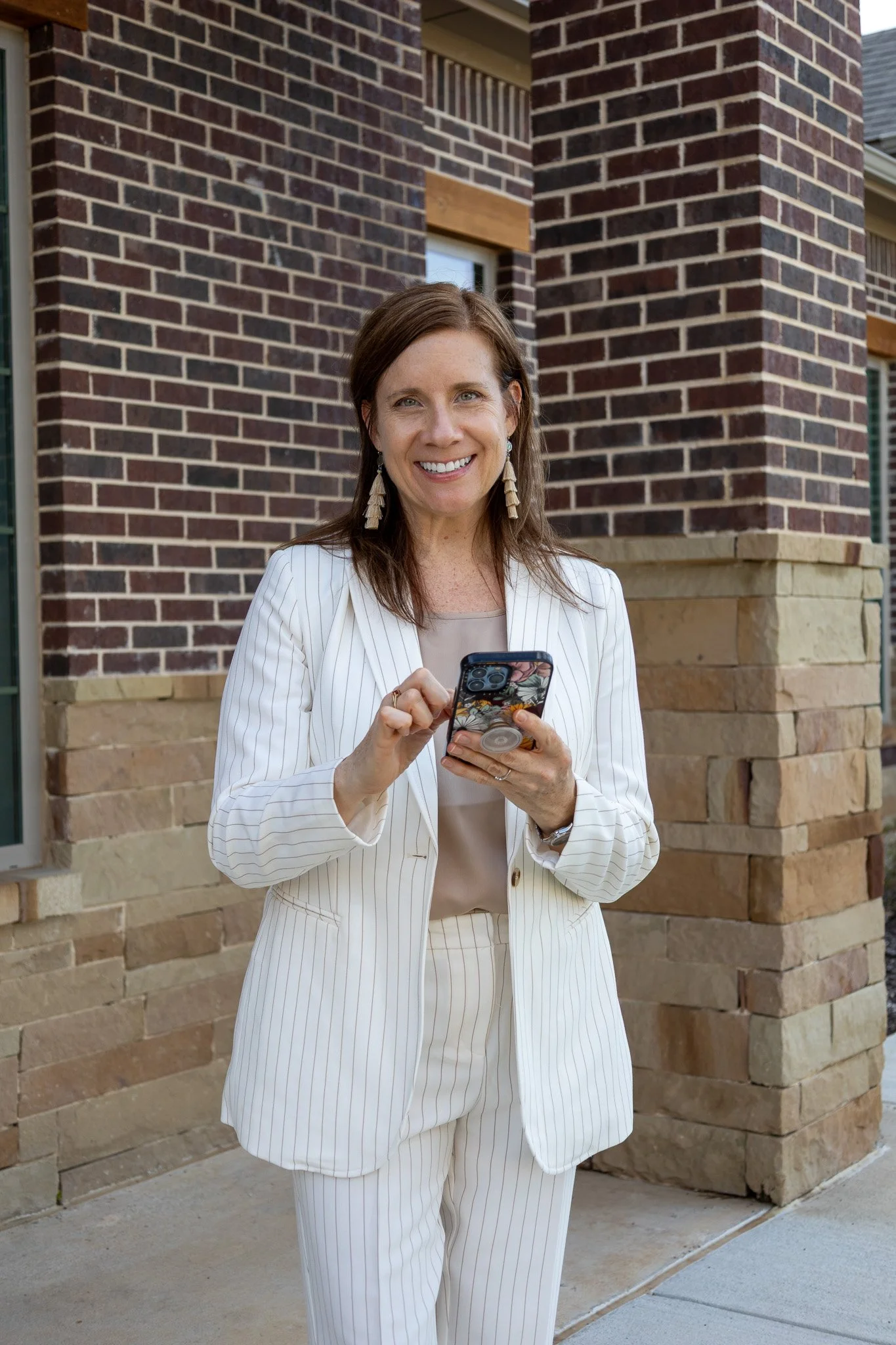 A woman dressed in a cream pinstriped suit stands outside in front of a brick building, smiling while using her smartphone.