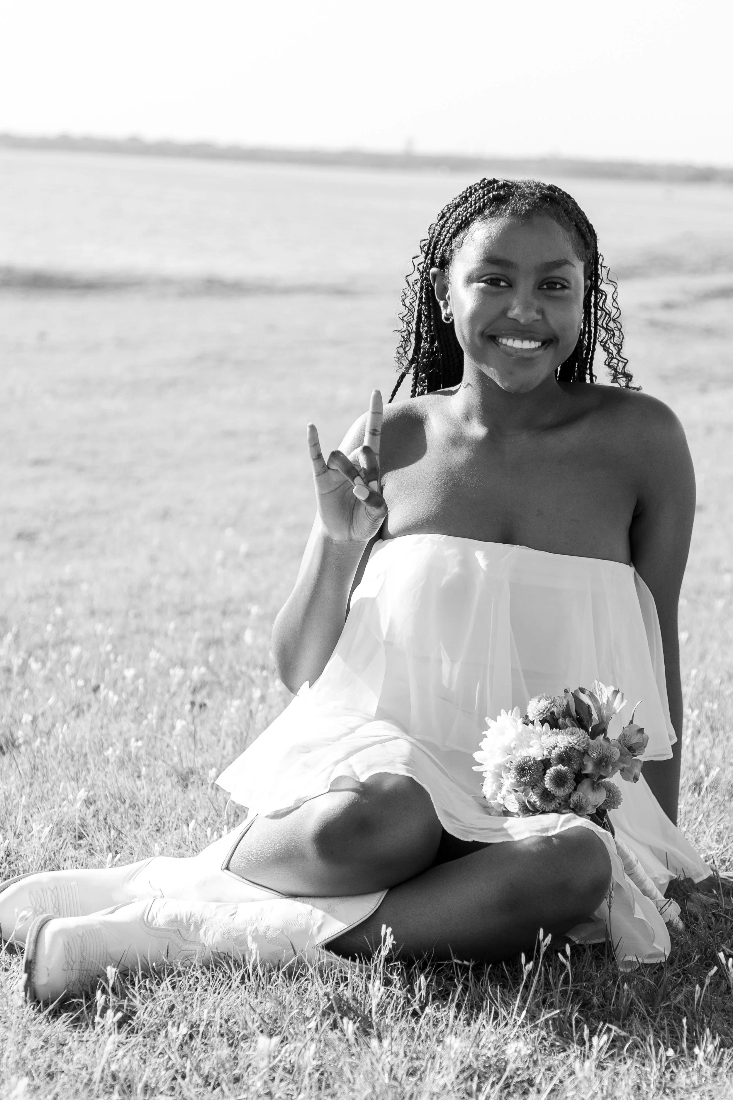 A smiling woman in a strapless dress sitting on the grass at the beach, making a peace sign with her right hand, holding a flower bouquet in her lap.