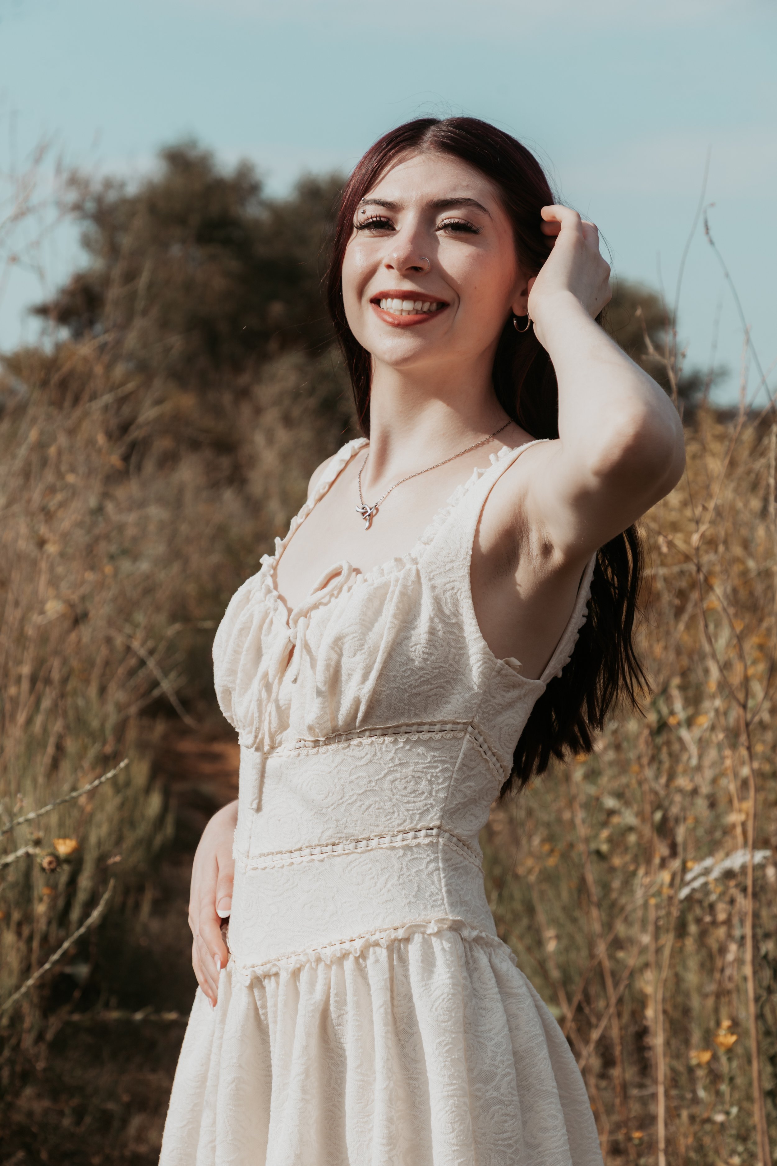 A young woman with dark hair and a nose piercing smiling outdoors in a field with dry grass, dressed in a light-colored, sleeveless, textured dress.
