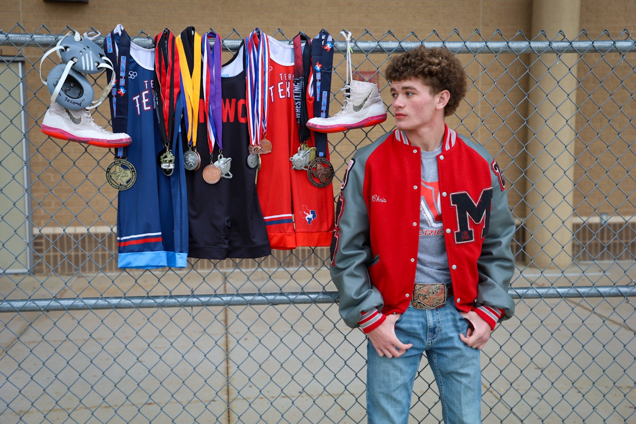 A young man with curly hair stands next to a fence displaying medals, athletic shoes, and sports jerseys. The medals hang from ribbons, and the jerseys are draped over the fence. The young man is wearing a red and gray varsity jacket and jeans.