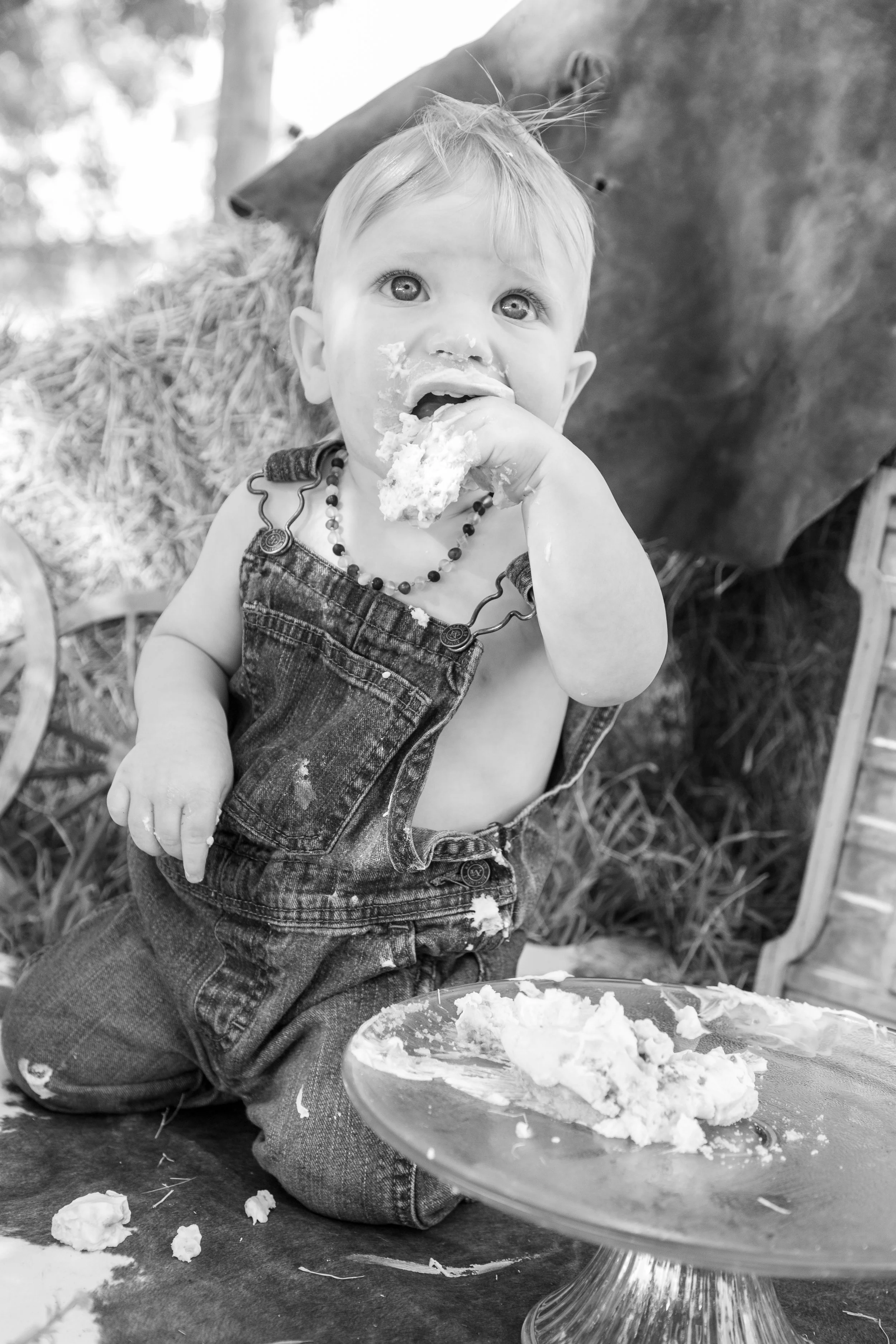 A young child with light-colored hair, dressed in denim overalls and a beaded necklace, sitting on the ground, eating cake with a messy face, surrounded by frosting and cake crumbs, with a cake stand in front of them.