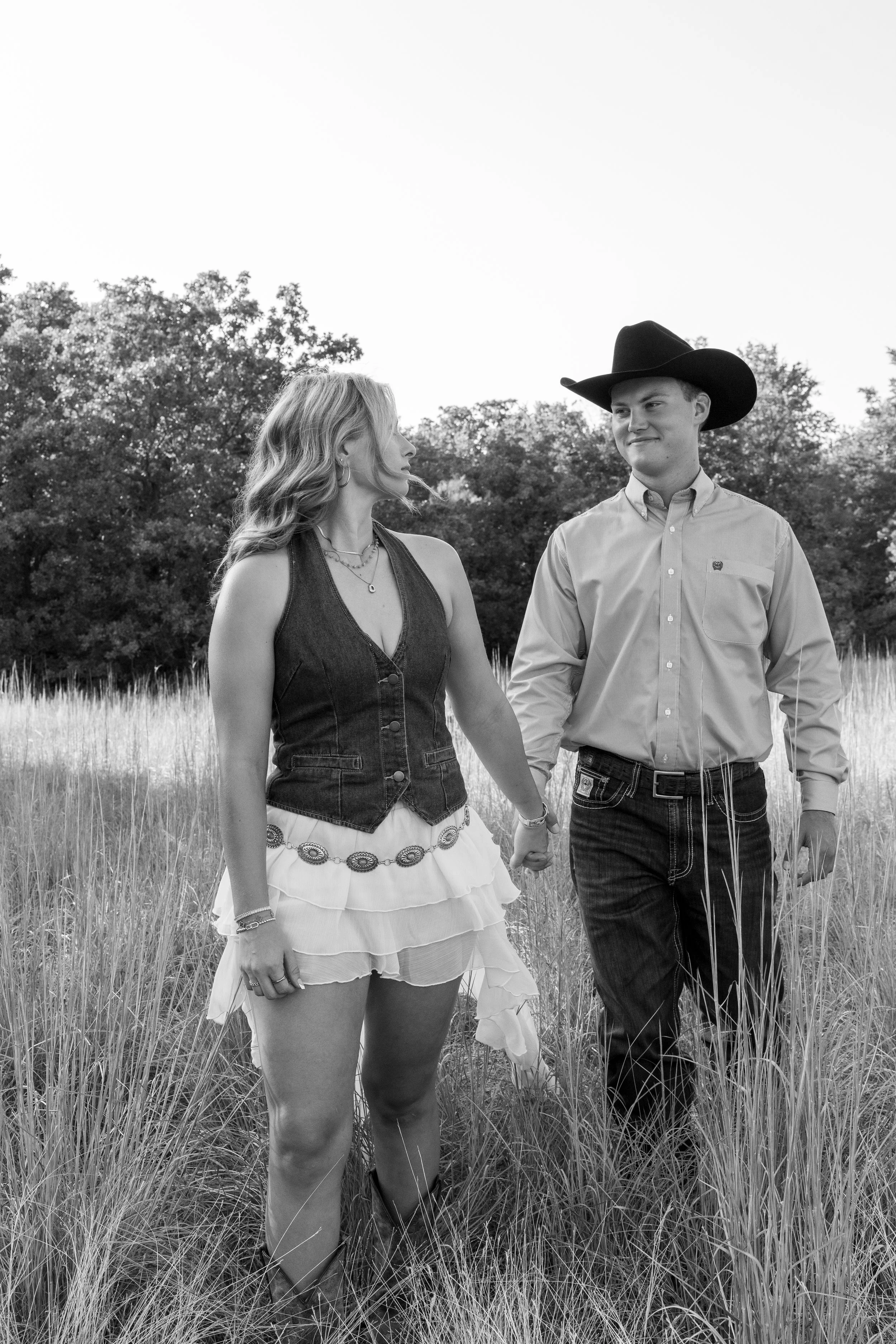 A couple holds hands and walks in a grassy field with trees in the background. The woman is wearing a sleeveless denim vest over a white, layered skirt, and has wavy hair. The man is wearing a cowboy hat, a long-sleeve button-up shirt, and dark jeans