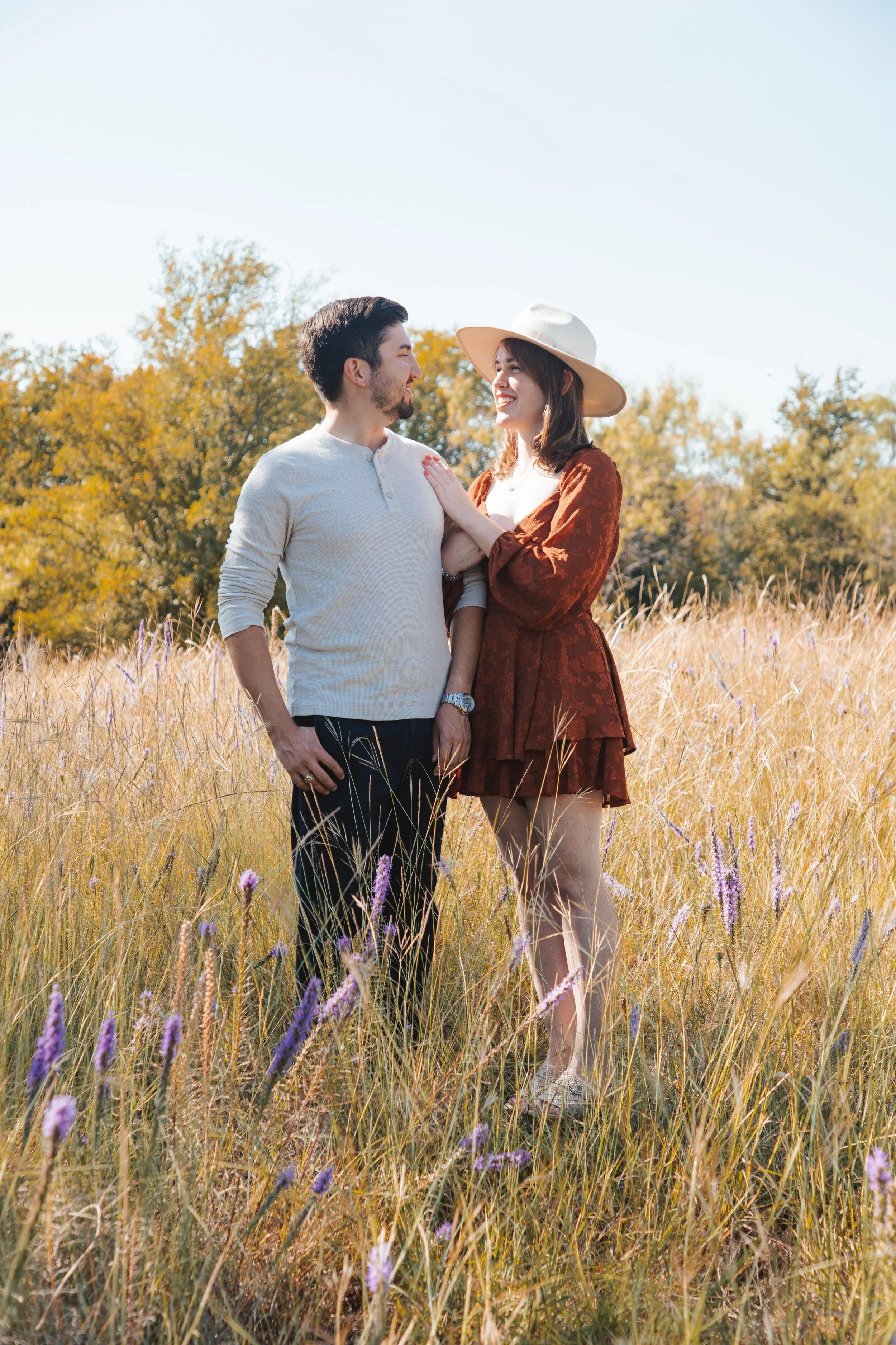 A man and woman standing in a field of tall grasses and purple flowers, looking at each other and smiling, with trees in the background and a clear blue sky.