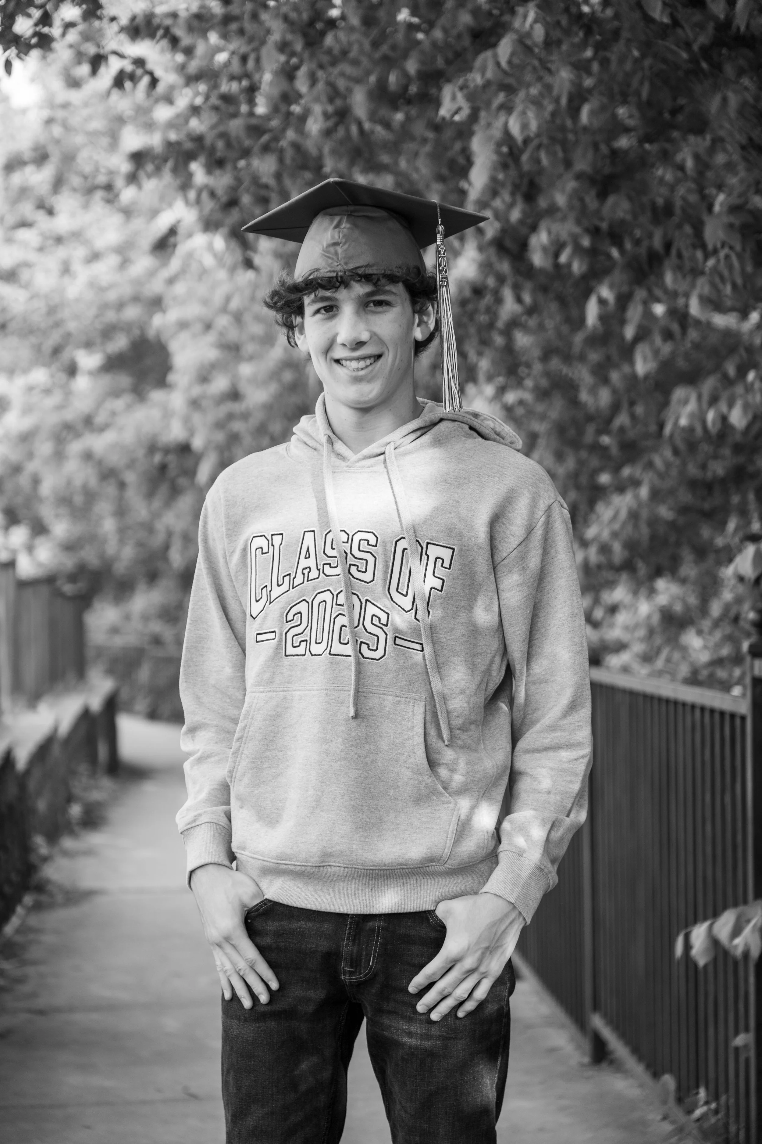 A young man wearing a graduation cap and hoodie, smiling outdoors on a pathway surrounded by trees.