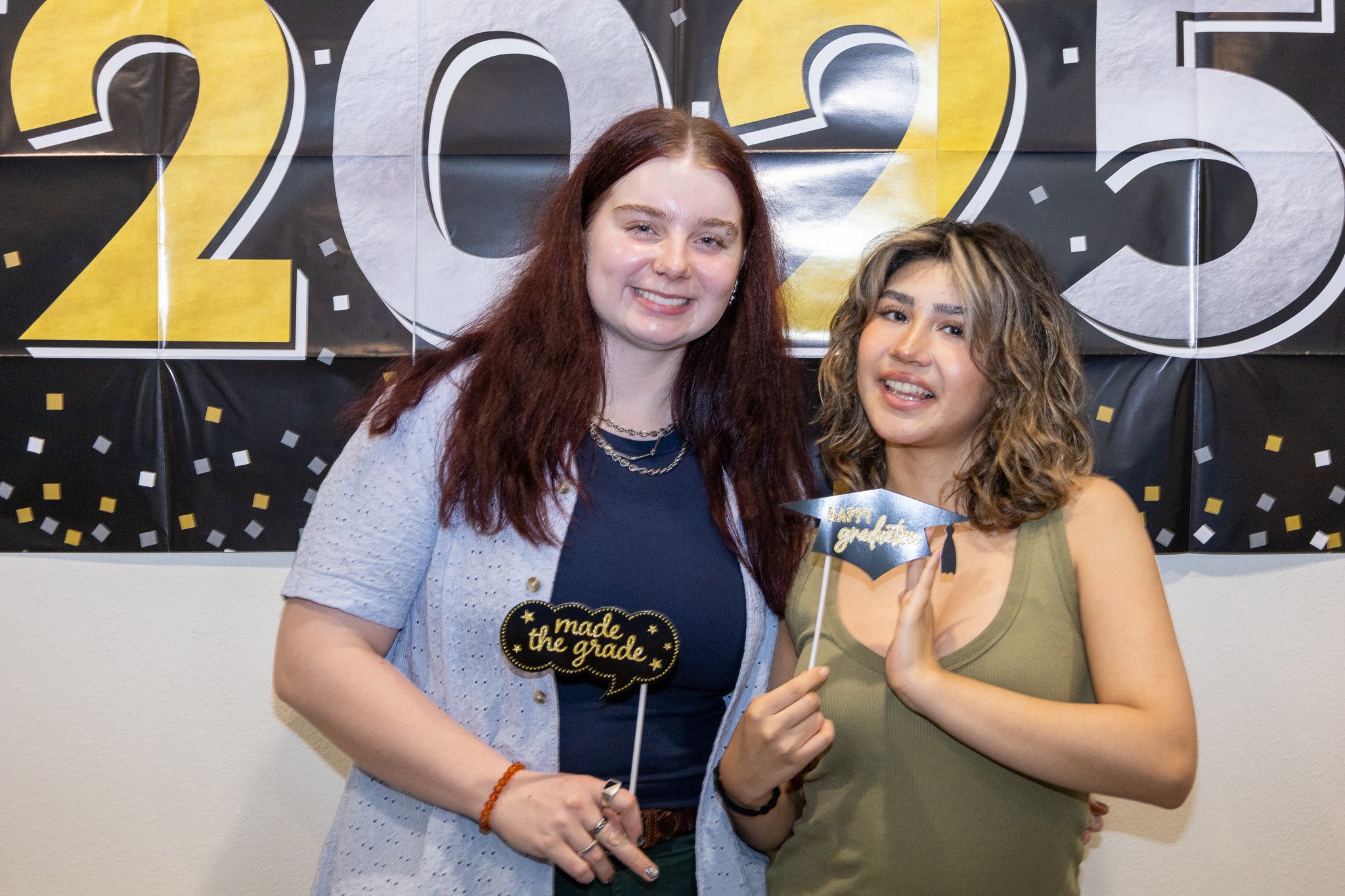 Two young women celebrating graduation, holding graduation-themed photo props, standing in front of a black and gold '2023' banner with confetti design.