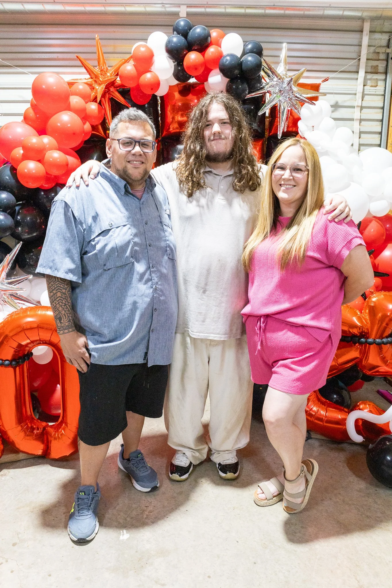 Three people standing in front of a balloon arrangement, smiling, with balloons in red, black, white, and silver colors. One man has glasses and a tattoo on his arm, a woman has long blonde hair, and the man in the middle has long curly hair.