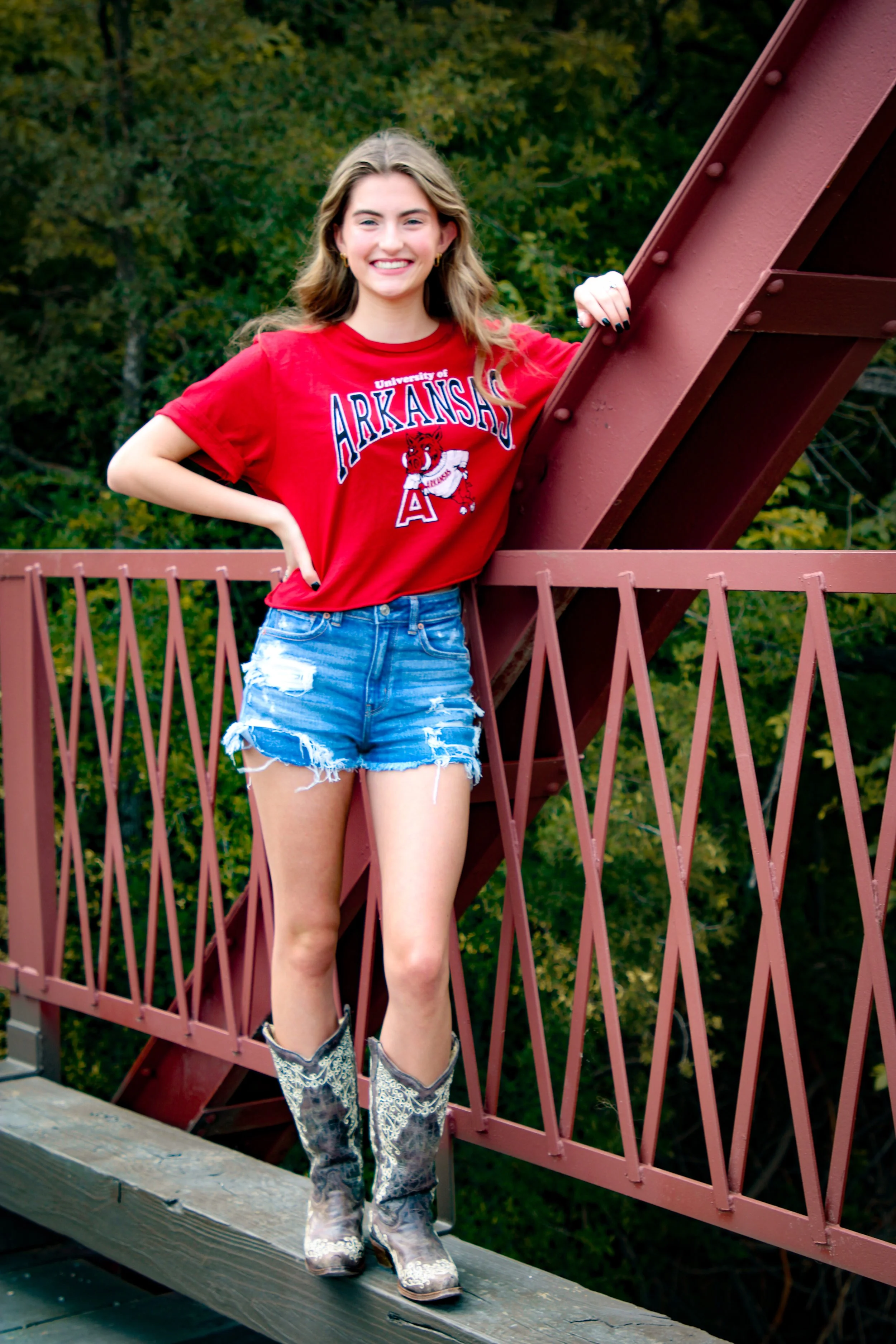 Young woman standing on a bridge wearing a red University of Arkansas t-shirt, distressed denim shorts, and cowboy boots, smiling at the camera with a green forest background.