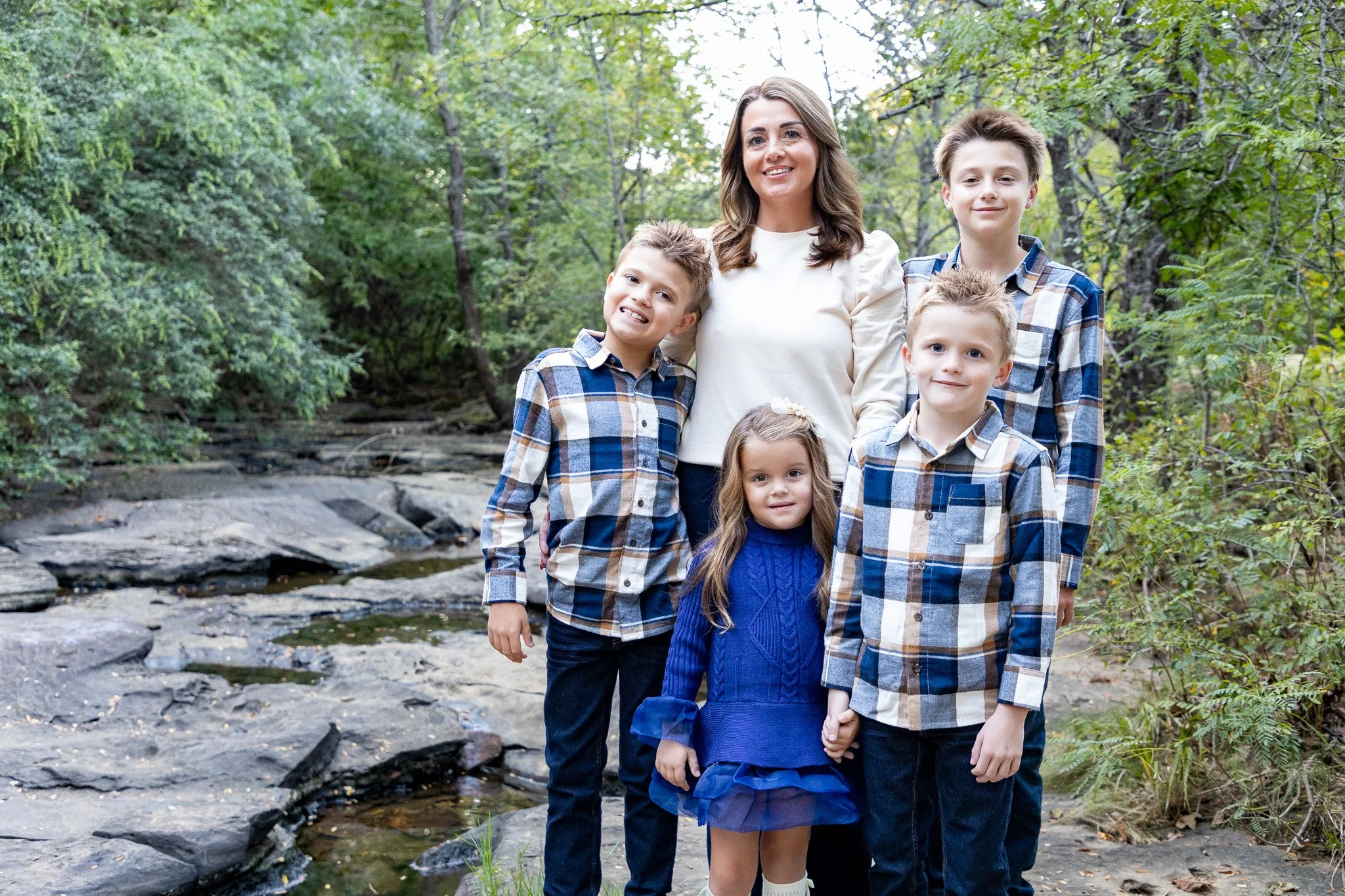 A woman and five children standing together outdoors near a creek, with trees and greenery in the background.
