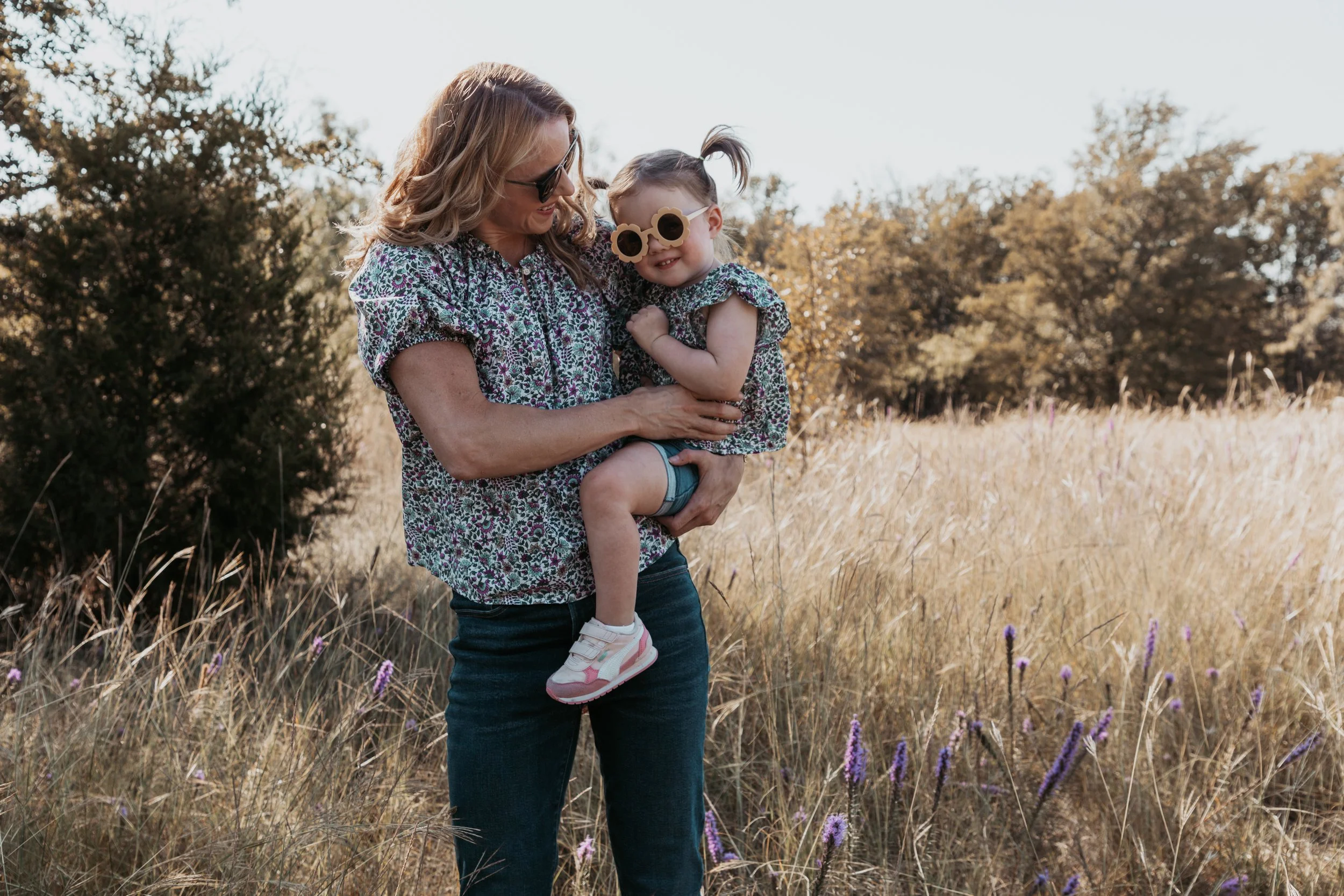 A woman holding a young girl in a field of tall grass and purple flowers, smiling and wearing sunglasses on a sunny day.