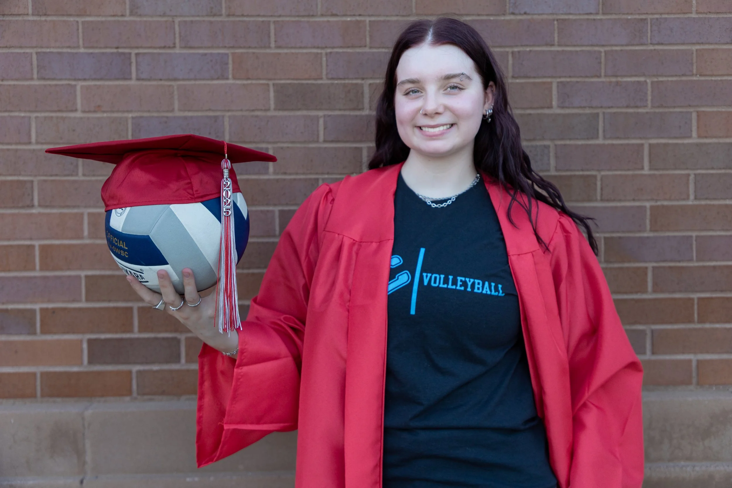 A young woman in a black volleyball t-shirt and red graduation gown holding a volleyball with a red graduation cap on it, standing in front of a brick wall.