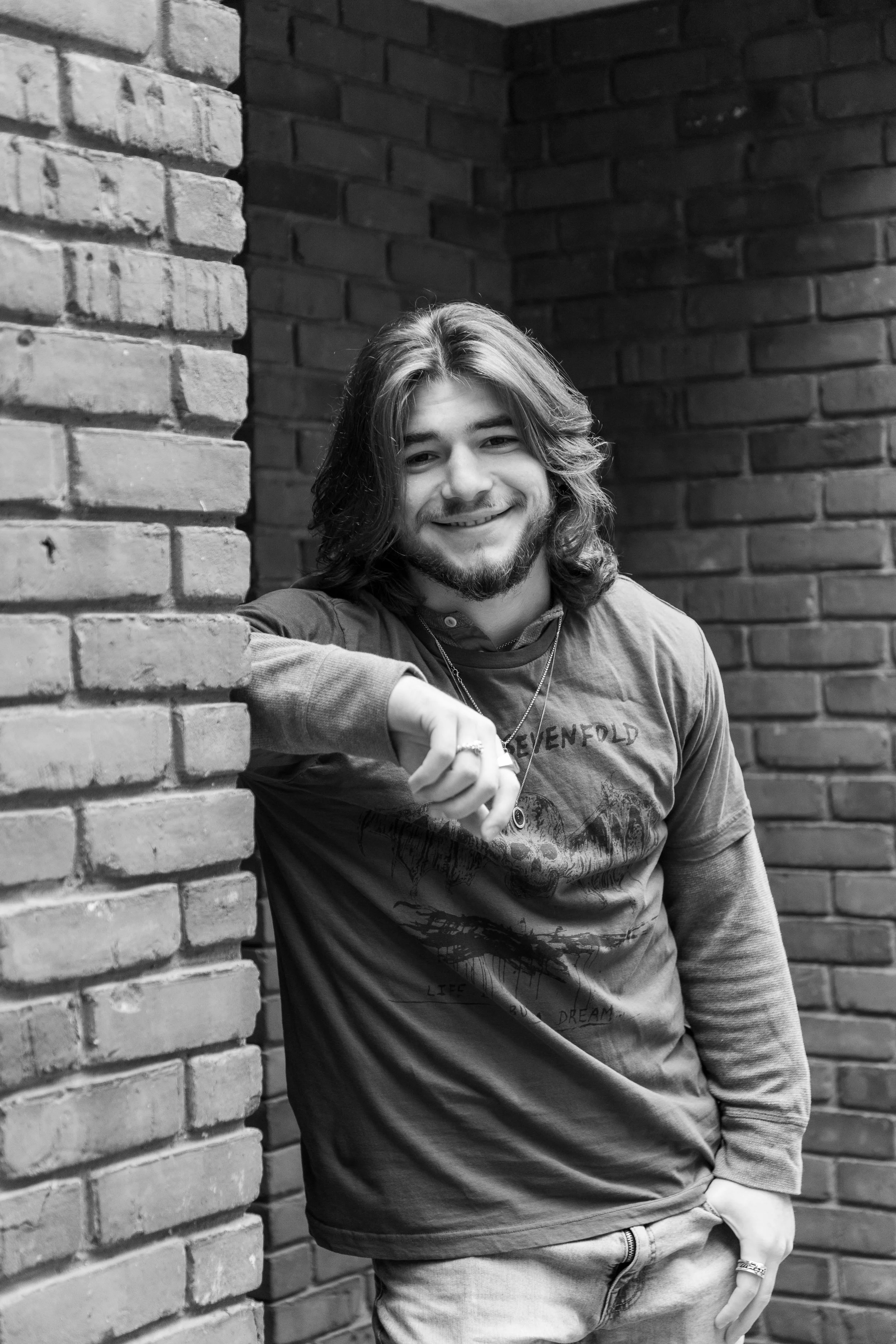 A young man with long hair and a beard smiling, leaning against a brick wall. He is wearing a graphic t-shirt and layered shirts, with rings and necklaces, in an indoor setting with brick walls.