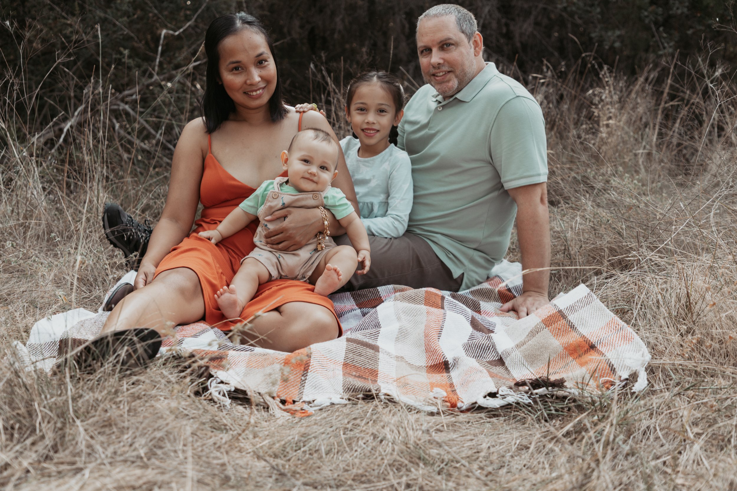 Family of four sitting on a blanket in a field, smiling at the camera.