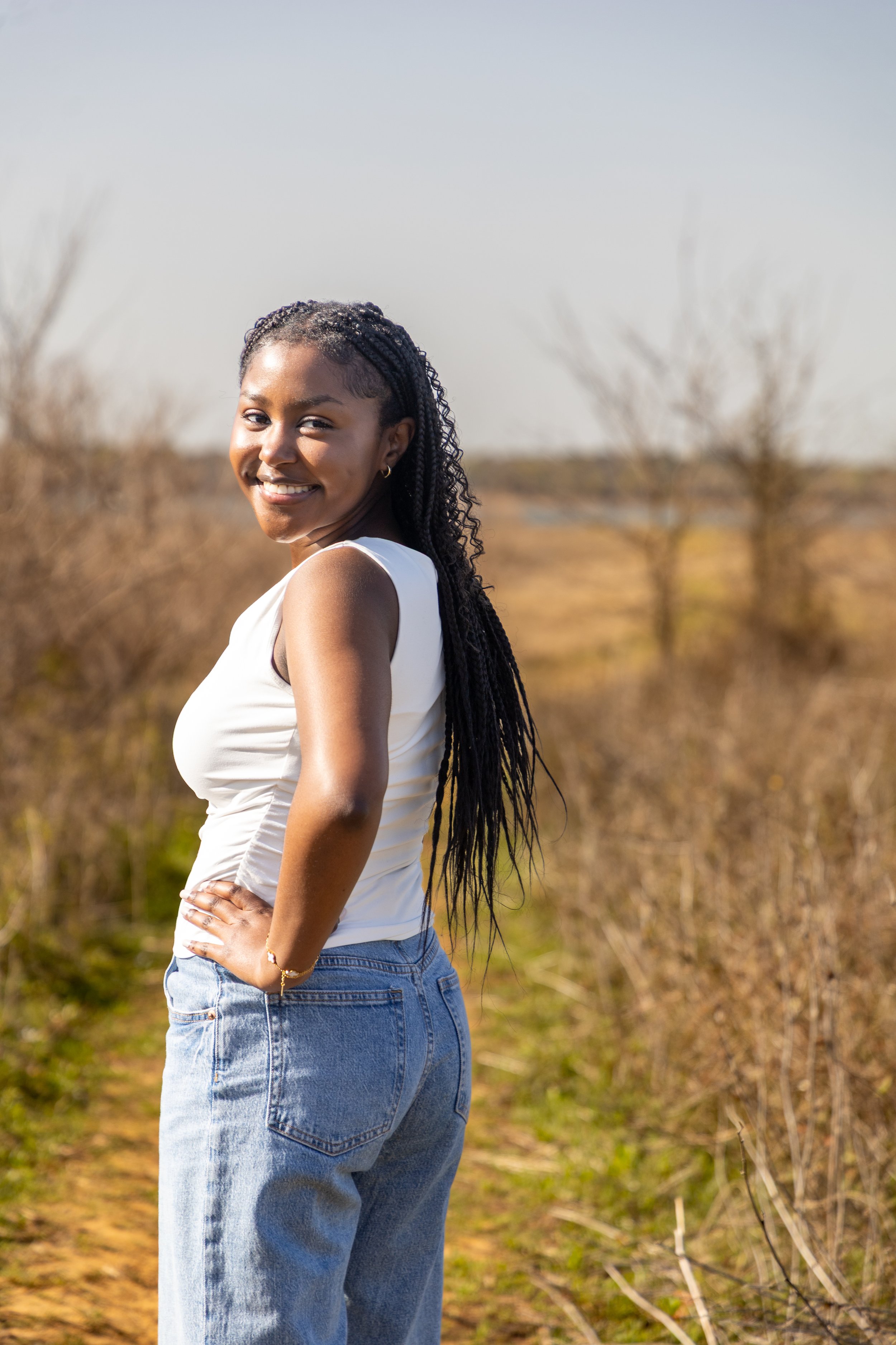 A young woman with long braided hair standing outdoors on a dirt path in a rural setting, smiling at the camera.