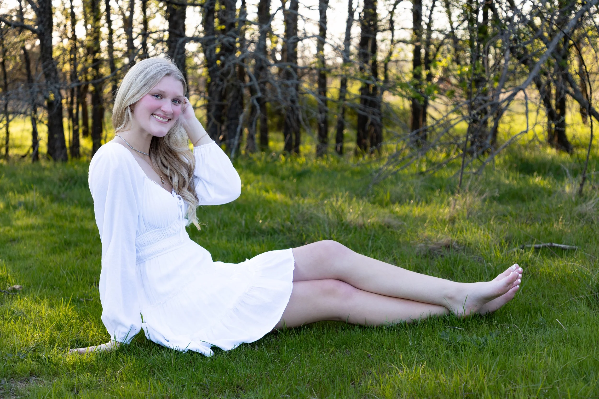 A young woman with long blonde hair, smiling and sitting on green grass in a forest, wearing a white dress.