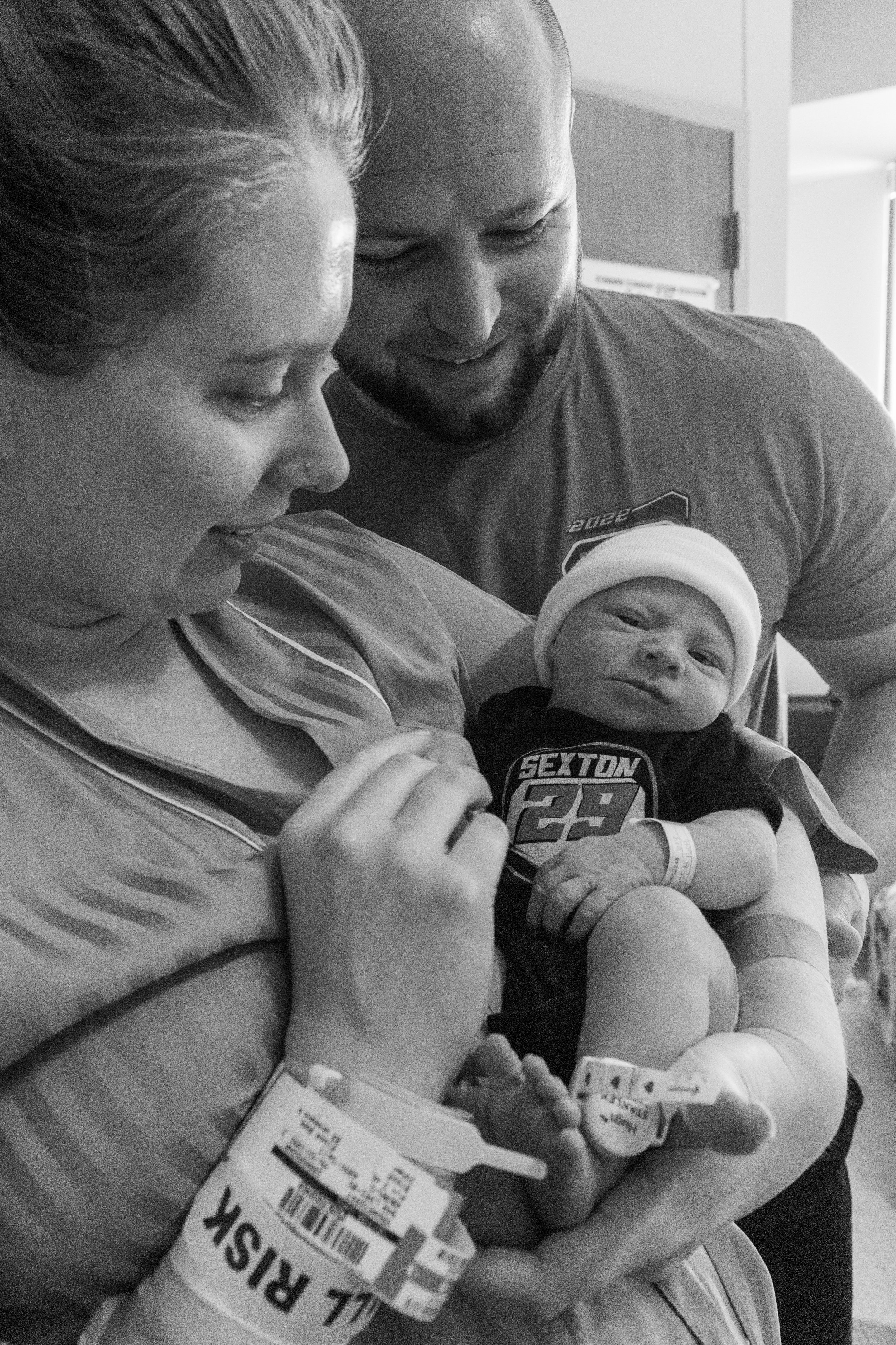 A woman holding a newborn baby in a hospital, with a man leaning in smiling, all three looking at the baby.