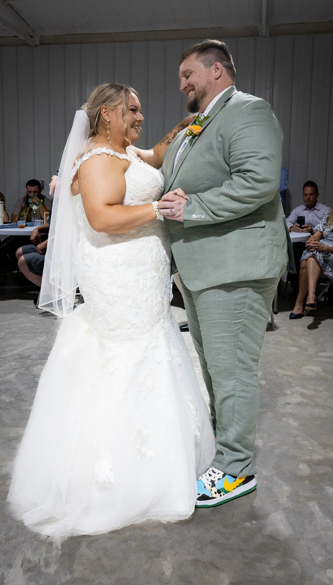 A bride and groom dance at their wedding reception, smiling and holding hands, with guests seated in the background.