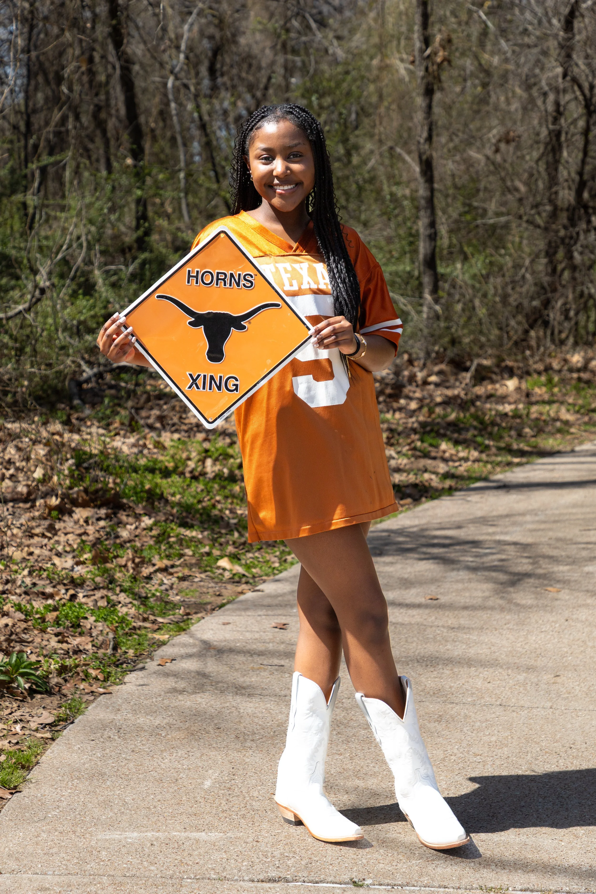 A young woman dressed in an orange University of Texas Longhorns football jersey and white cowboy boots holding a warning sign that says "Horns X ing" with a longhorn cattle head silhouette, standing on a sidewalk outdoors with trees in the backgroun