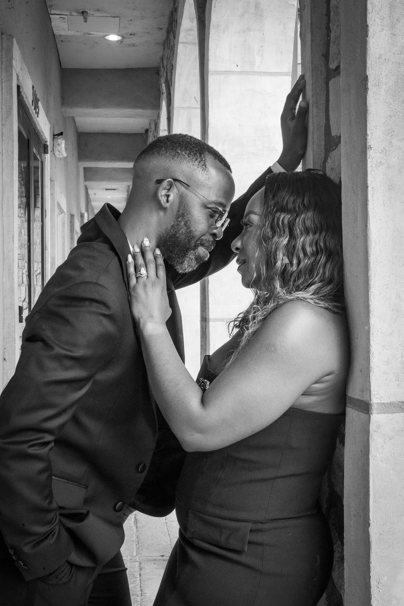 A black and white photo of a man and woman indoors, standing close with foreheads touching, gazing into each other's eyes. The woman has her hands on the man's chest and neck, and the man has one hand on the wall and the other near his pocket.