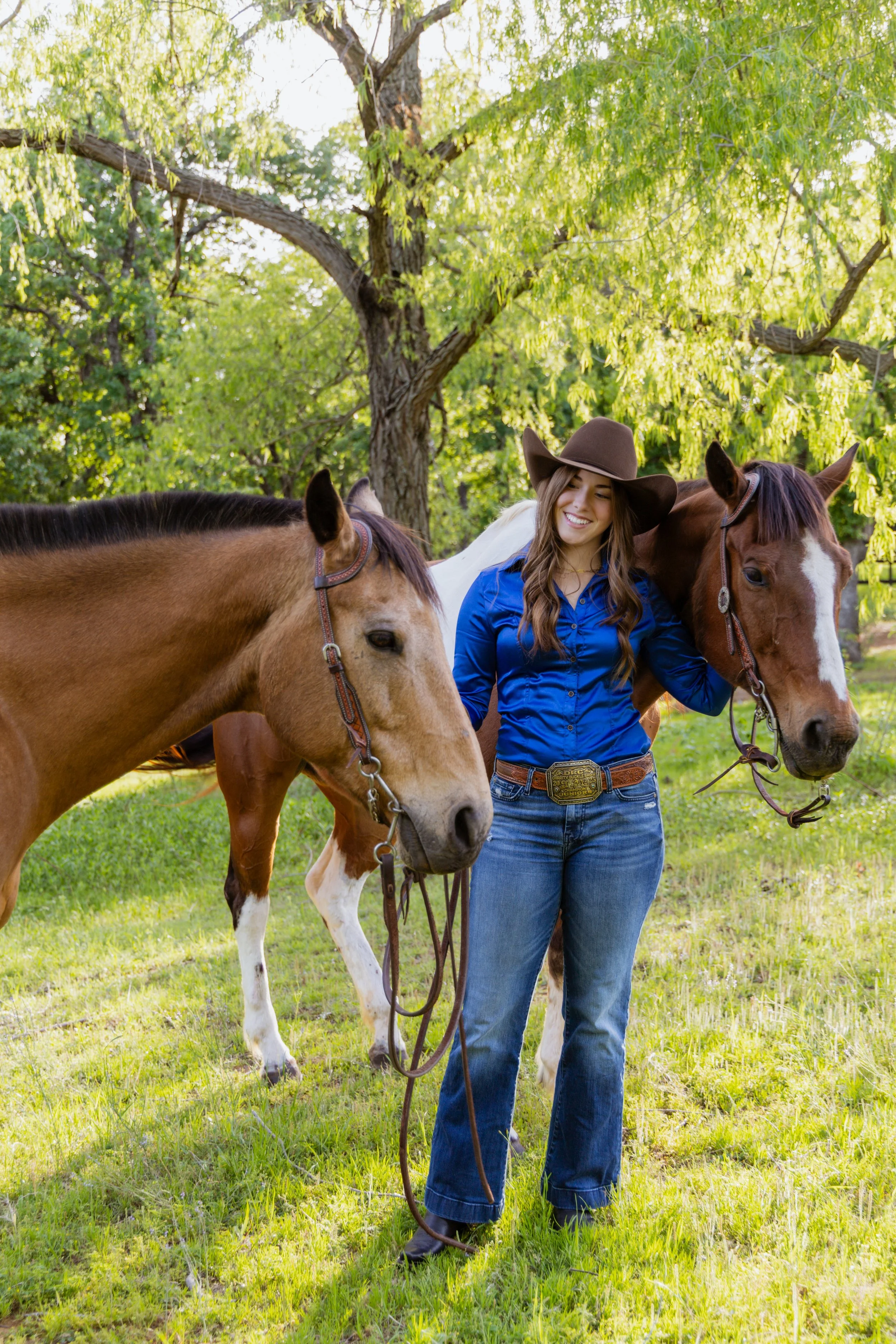 A young woman in a blue shirt and jeans standing on green grass, smiling, holding the reins of two brown horses with white markings on their faces under a large tree with green leaves.