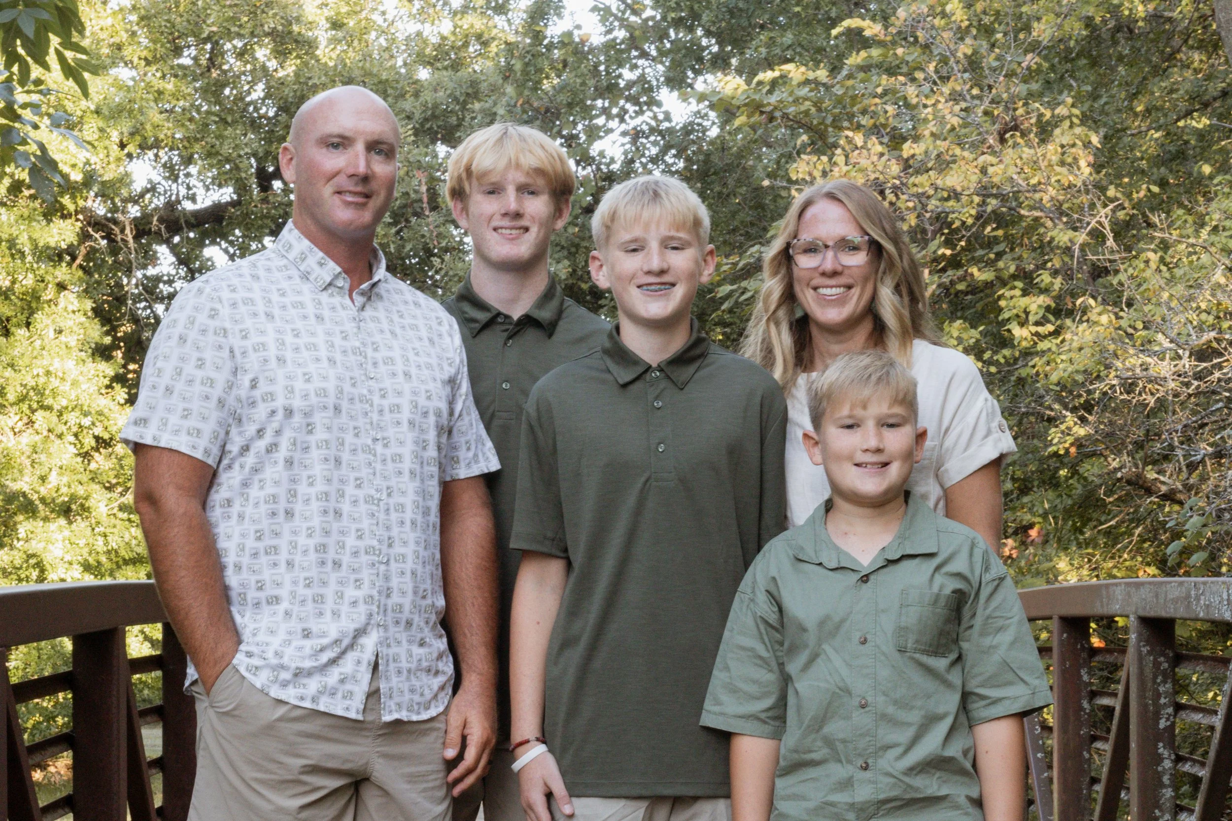 A family of six posing outdoors on a wooden bridge surrounded by green trees. The family includes a bald man wearing a white patterned shirt, a blonde woman with glasses wearing a light-colored shirt, and four boys in green shirts, all smiling.