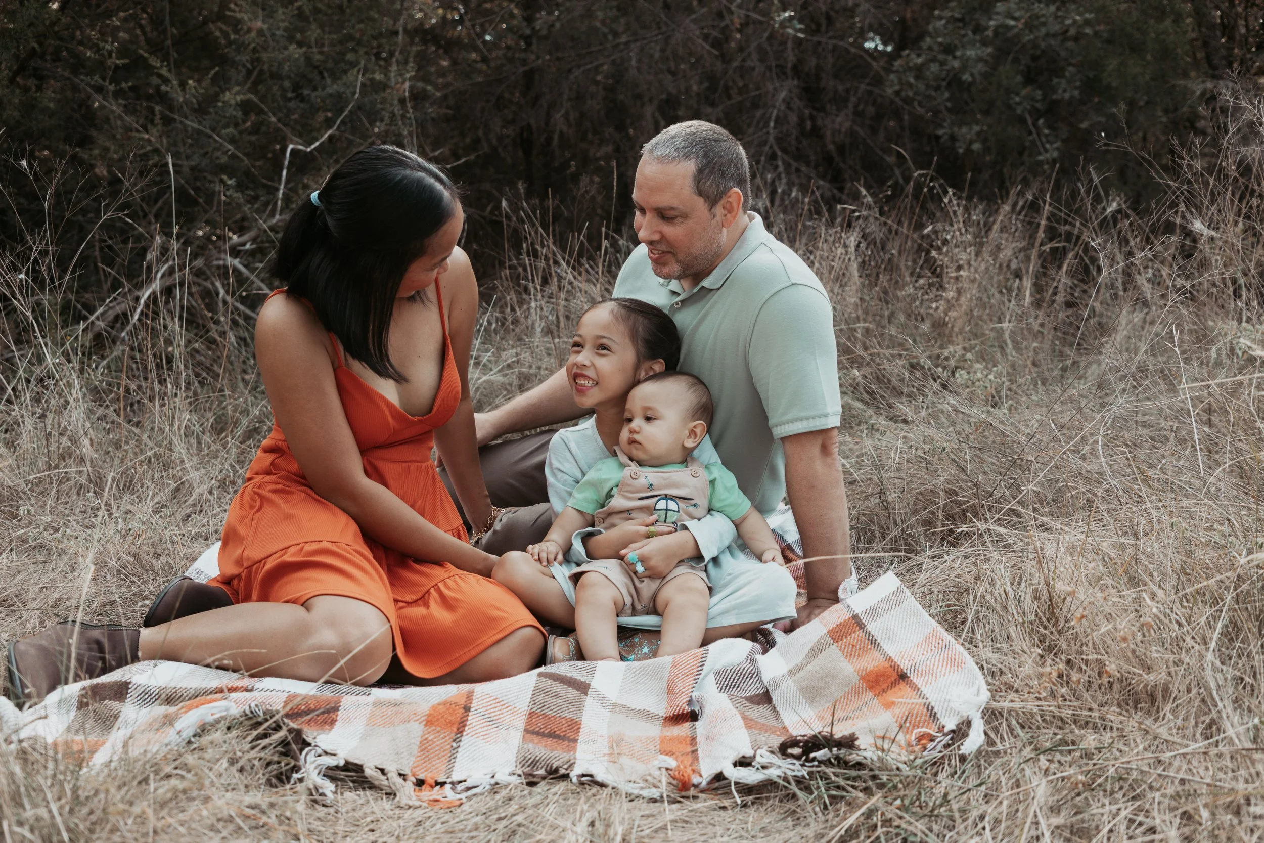 A family of four sitting on a picnic blanket in a field with dry grass and trees in the background. The mother, father, and two children are smiling and interacting.