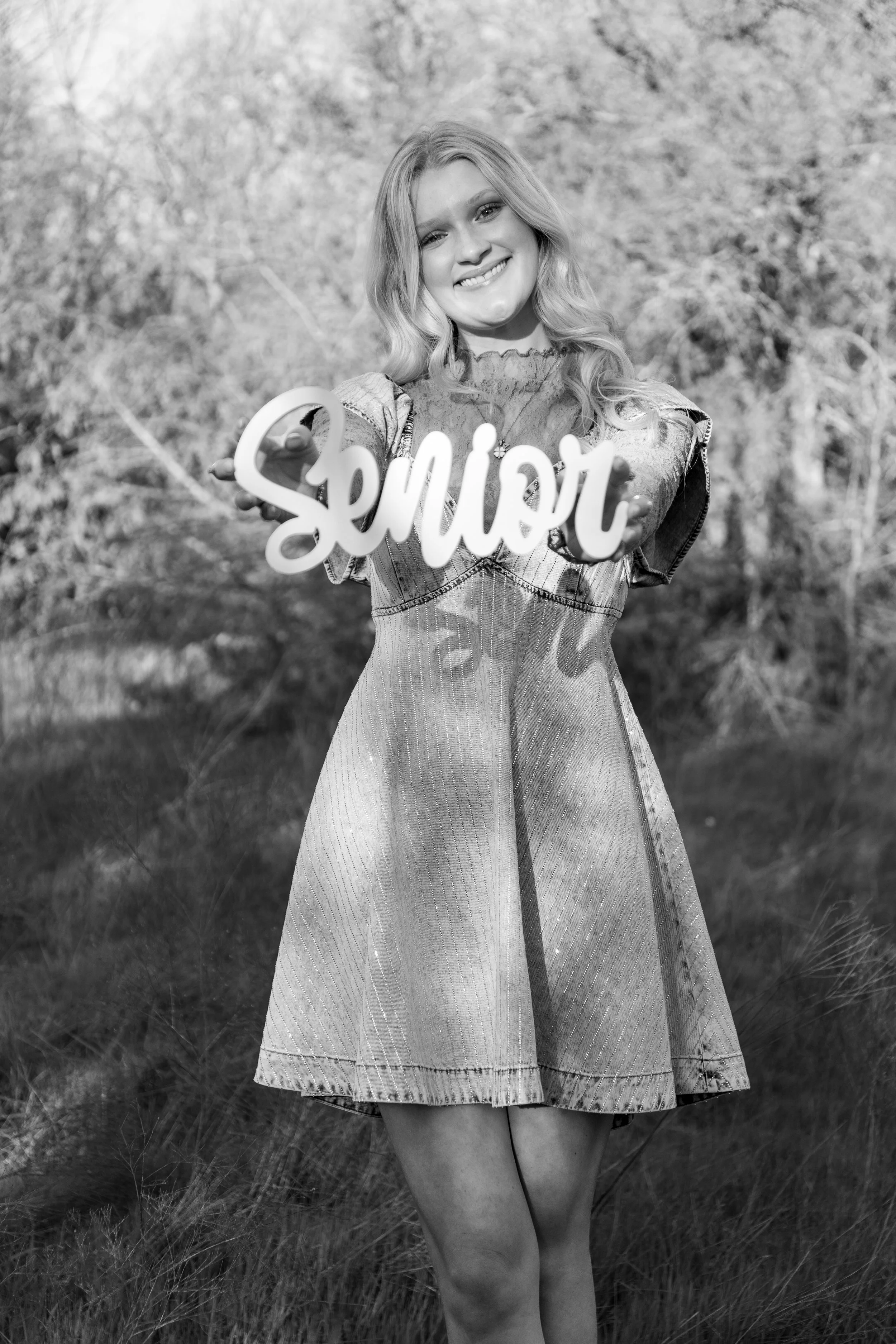 A young woman smiling outdoors, holding a sign that says "Senior," in a grassy area with trees in the background.