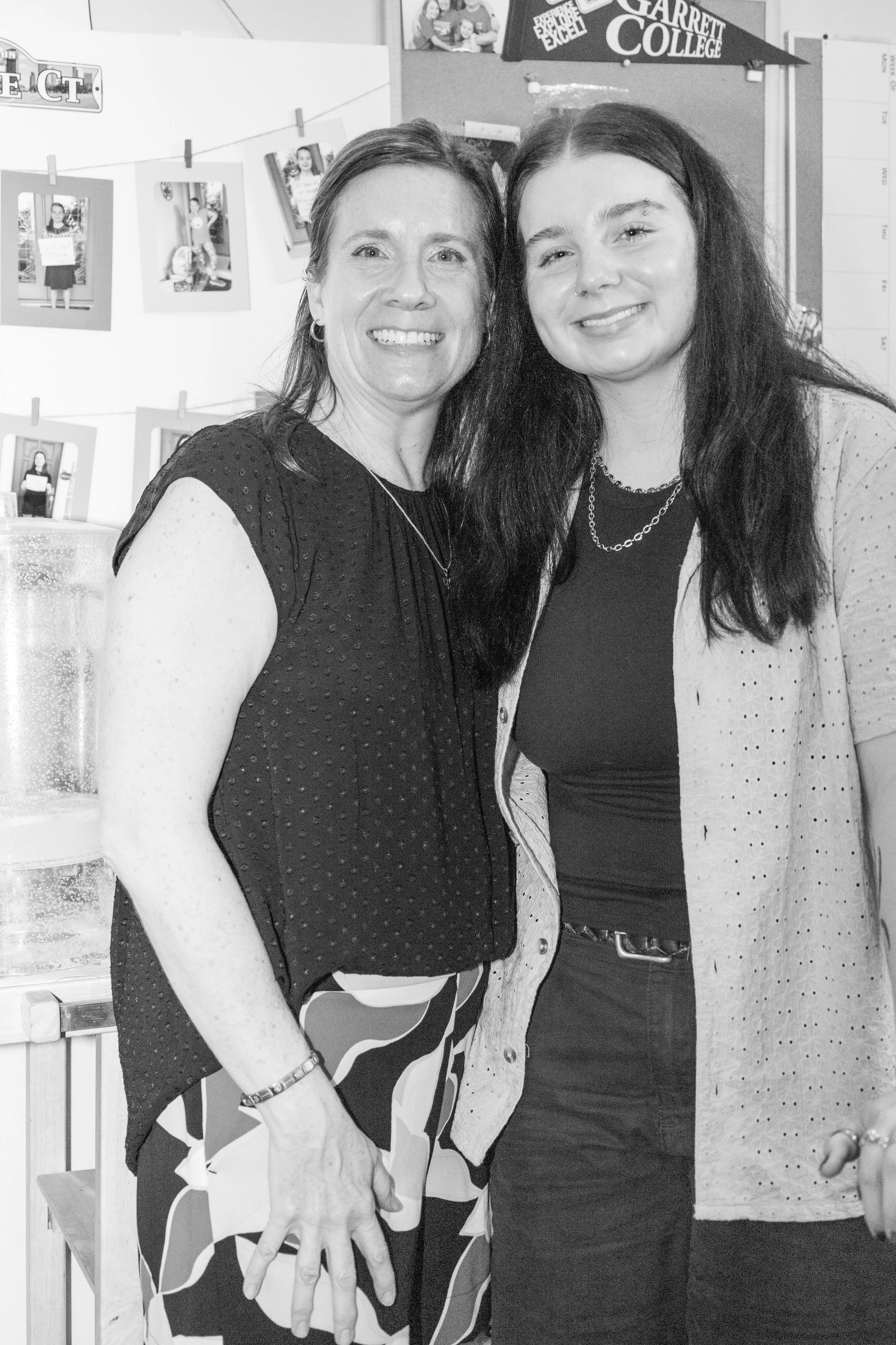 Two women smiling and standing close together indoors, with photos hanging on a display board behind them.