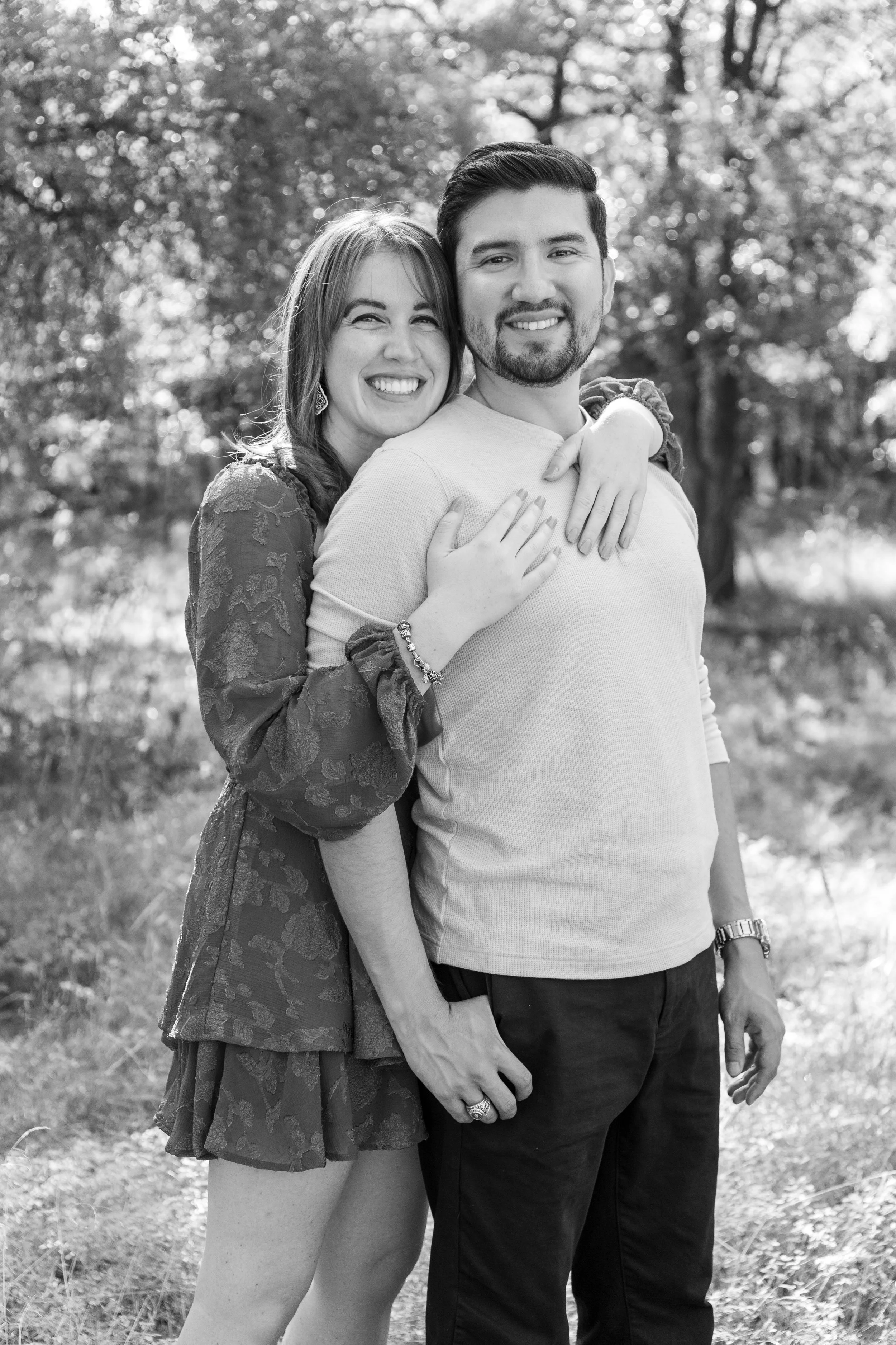 Black and white photo of a smiling woman hugging a man outdoors with trees in the background.