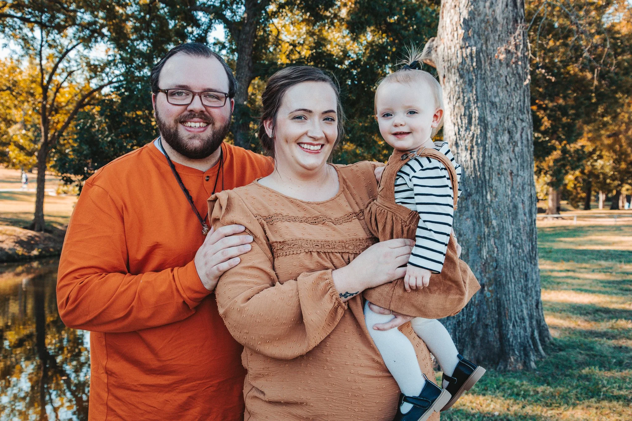 A family of three, including a man, woman, and young girl, smiling and posing outdoors in a park with trees and a pond in the background during autumn.