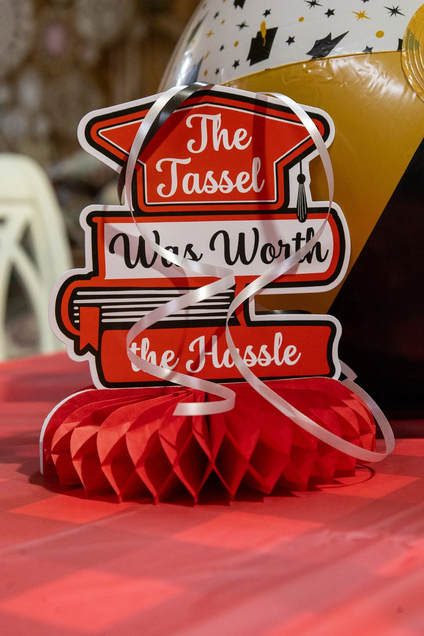 Decorative sign that says, 'The Tassel Was Worth the Hassle,' placed on red honeycomb paper at a party table.