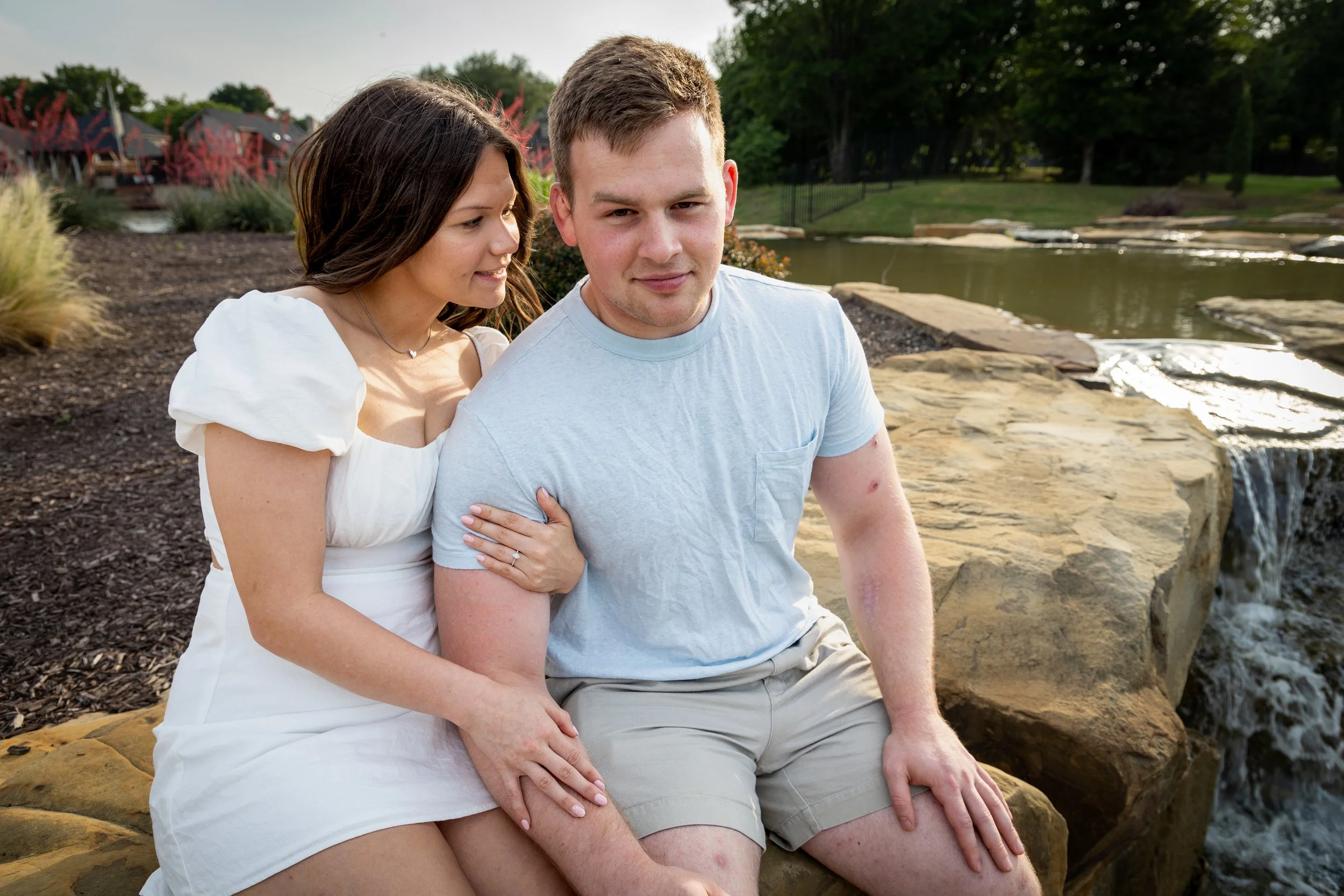 A woman in a white dress and a man in a light blue T-shirt sitting on rocks near a small waterfall, with greenery and houses in the background.