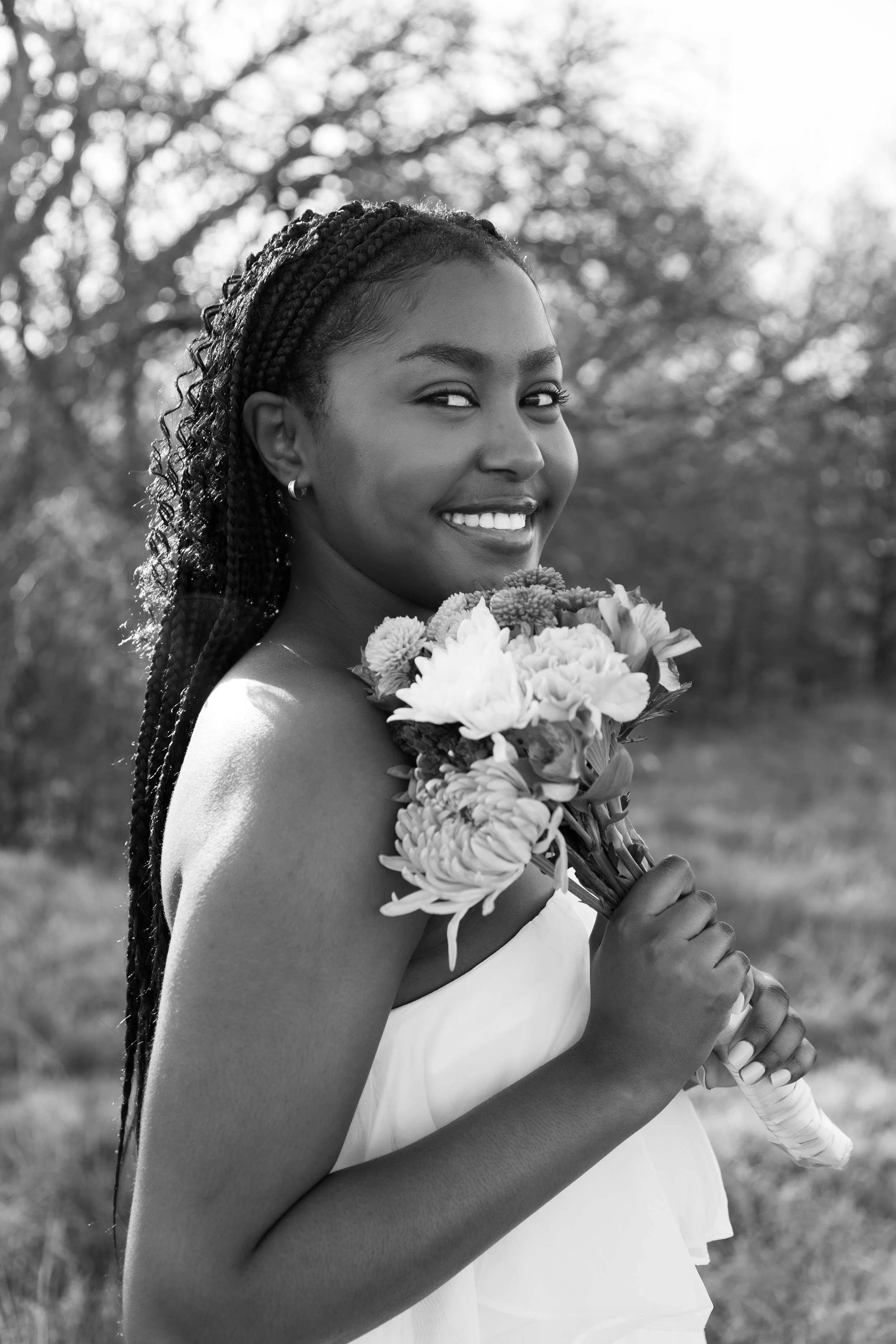 Smiling woman in a white dress holding a bouquet of flowers outdoors, with trees in the background, in black and white.