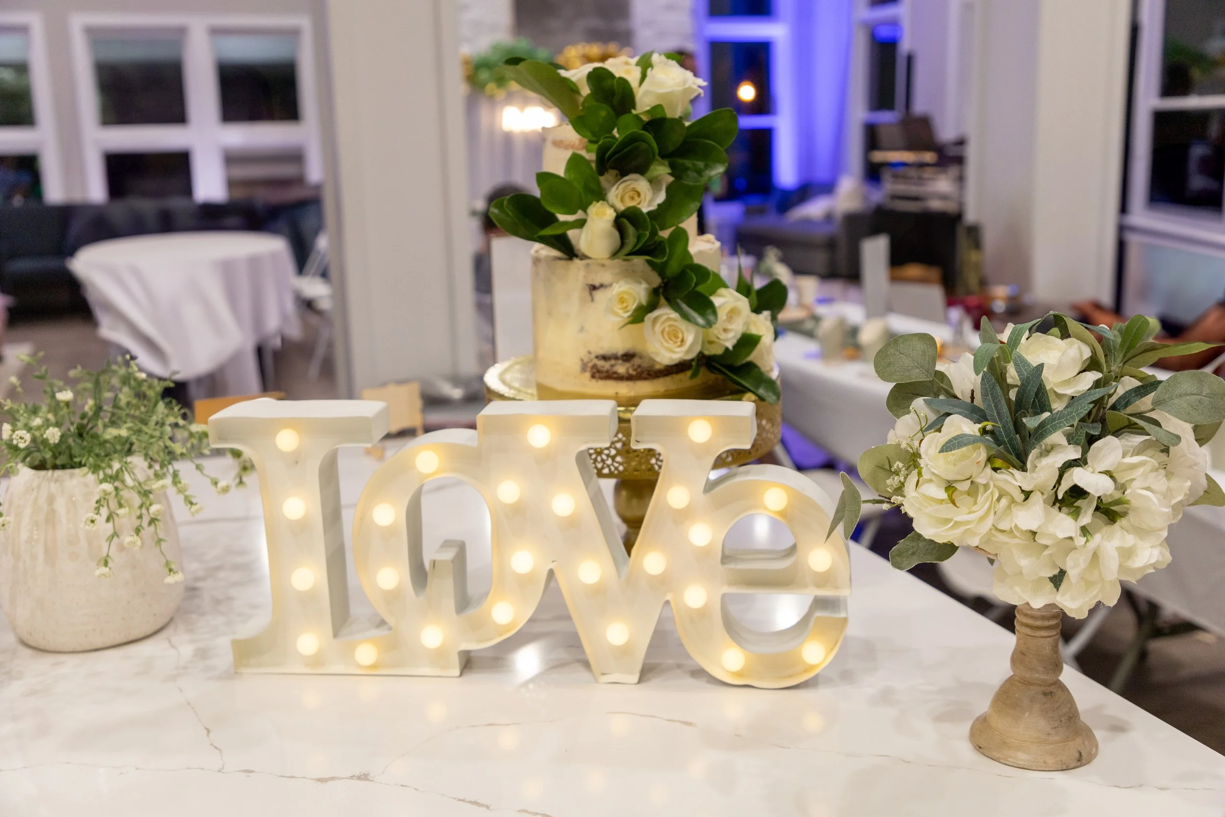Decorative table arrangement with a love sign with lights, a cake with white roses and greenery, and floral arrangements in pots, set in a festively decorated indoor space.