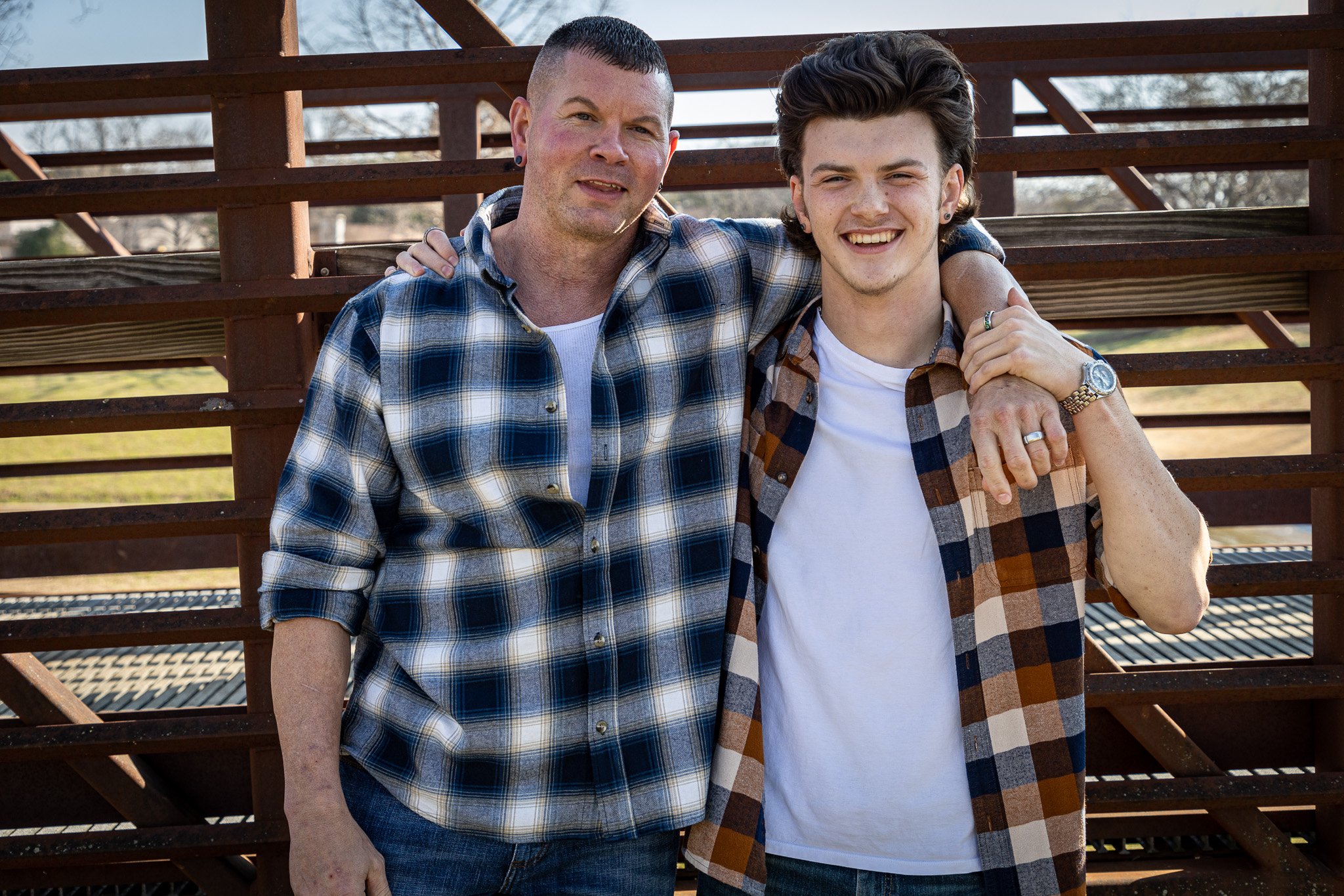 A father and son standing close together outdoors, smiling with arms around each other's shoulders, in front of a wooden staircase with a rural landscape in the background.