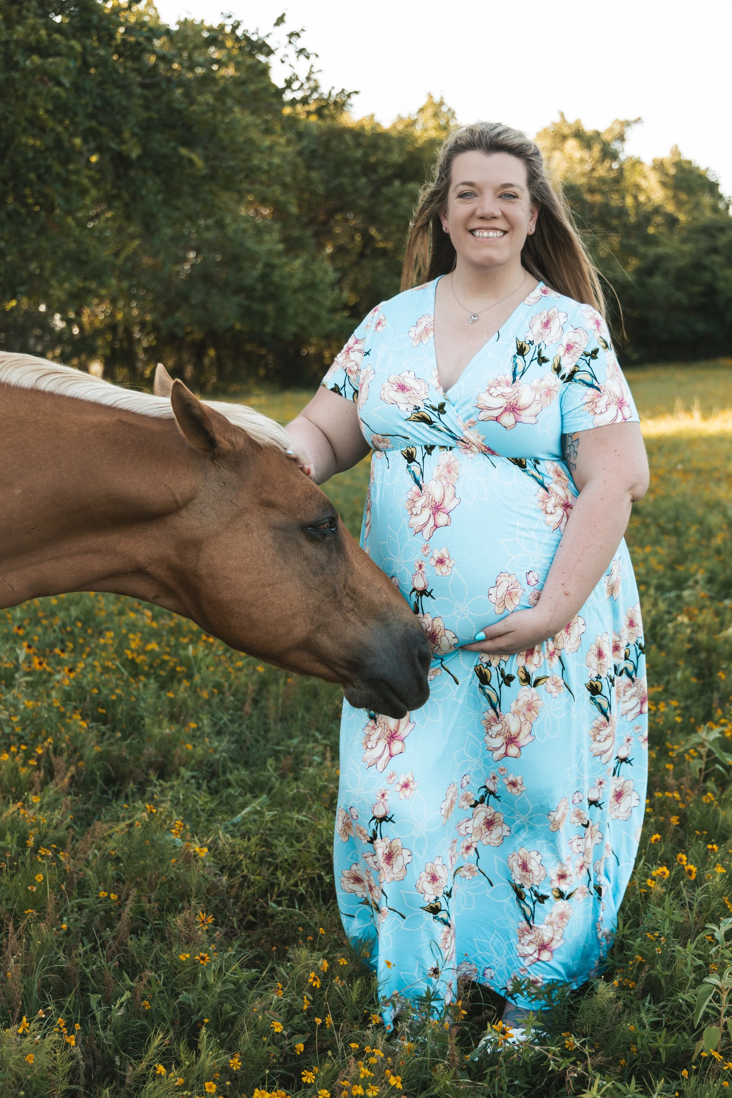 A woman in a blue floral dress stands in a field of yellow flowers, holding the reins of a brown horse, with green trees in the background, during sunset.