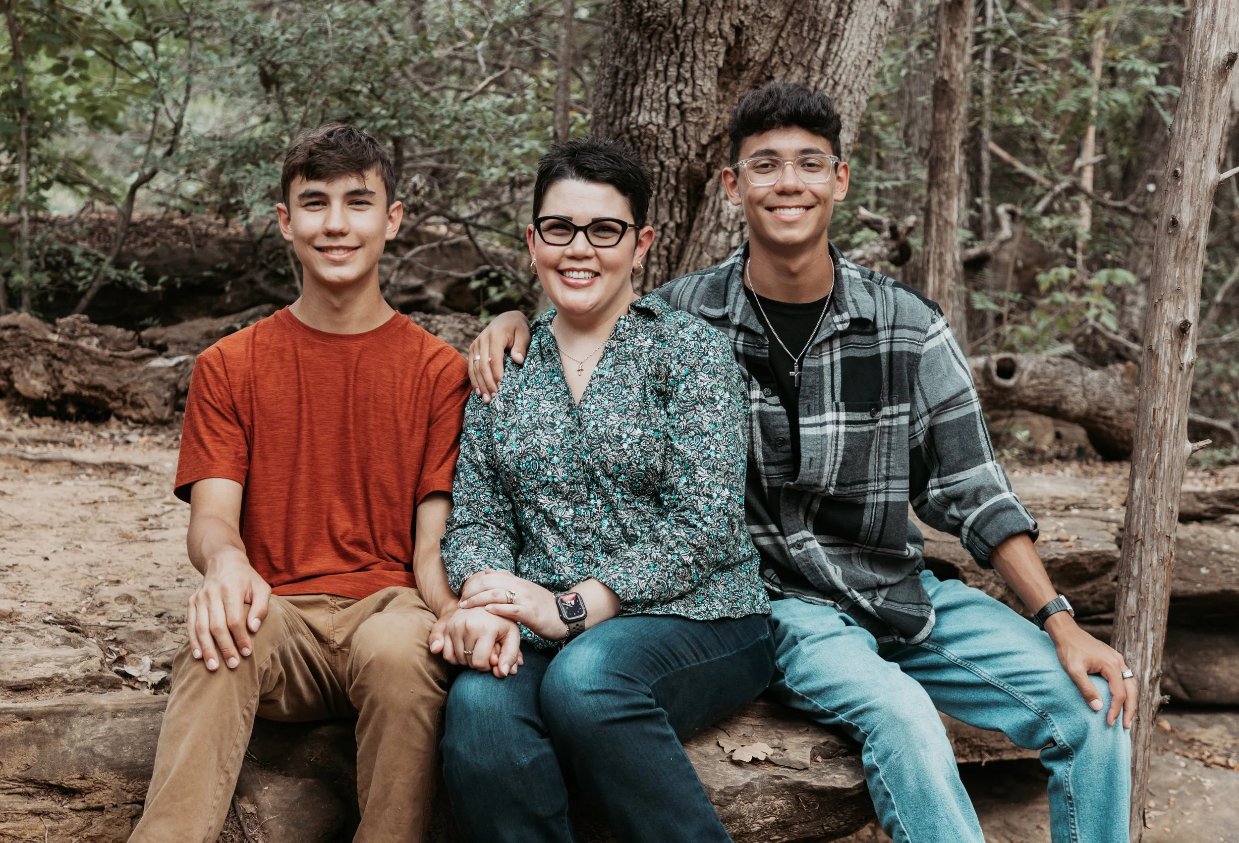 A woman with short black hair and glasses sitting between two teenage boys with short dark hair in a forest, all smiling at the camera.