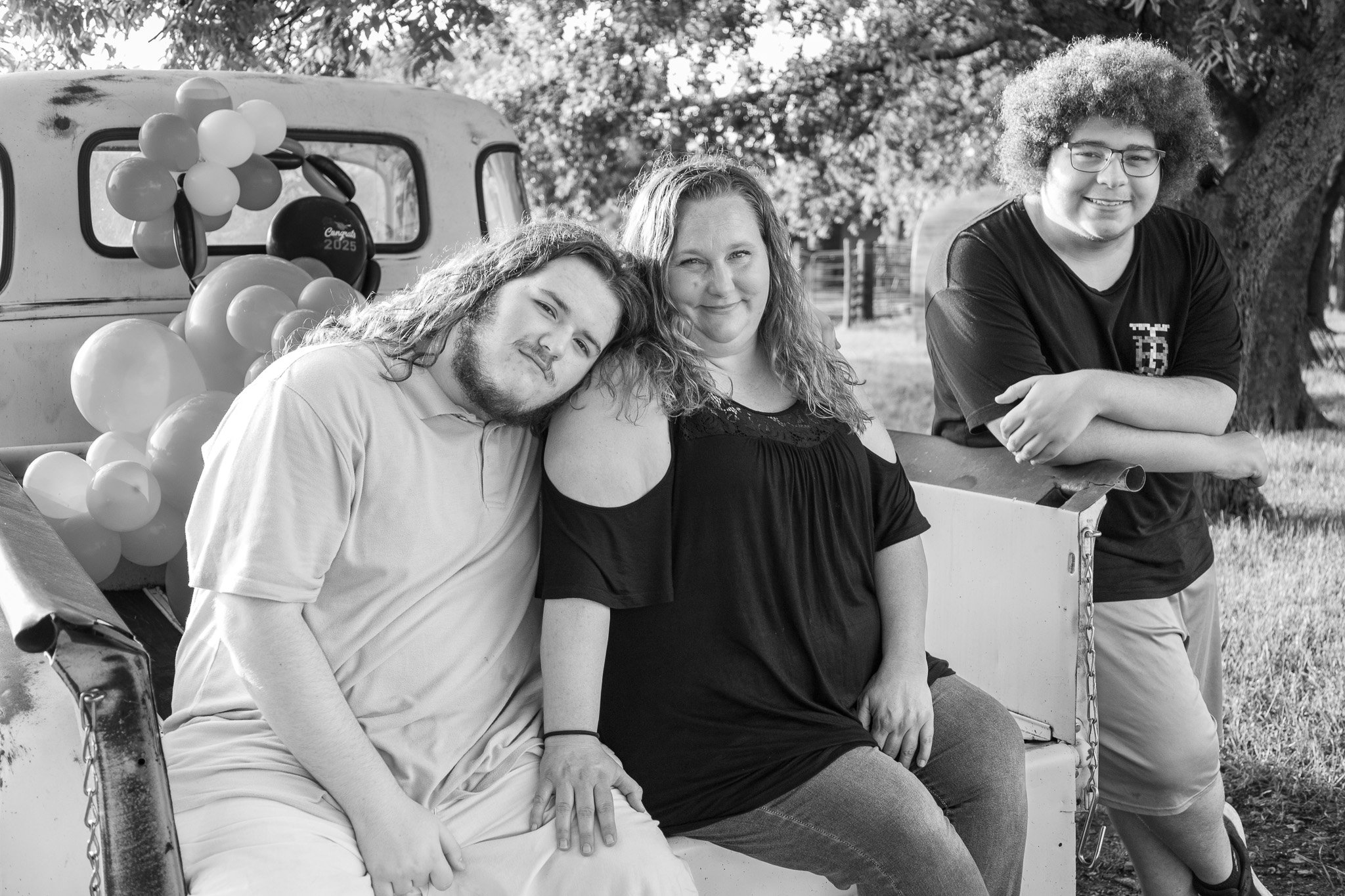 A black-and-white photo of three people sitting and leaning on an old pickup truck with balloons attached to it. They are smiling, outdoors, with trees and a fence in the background.