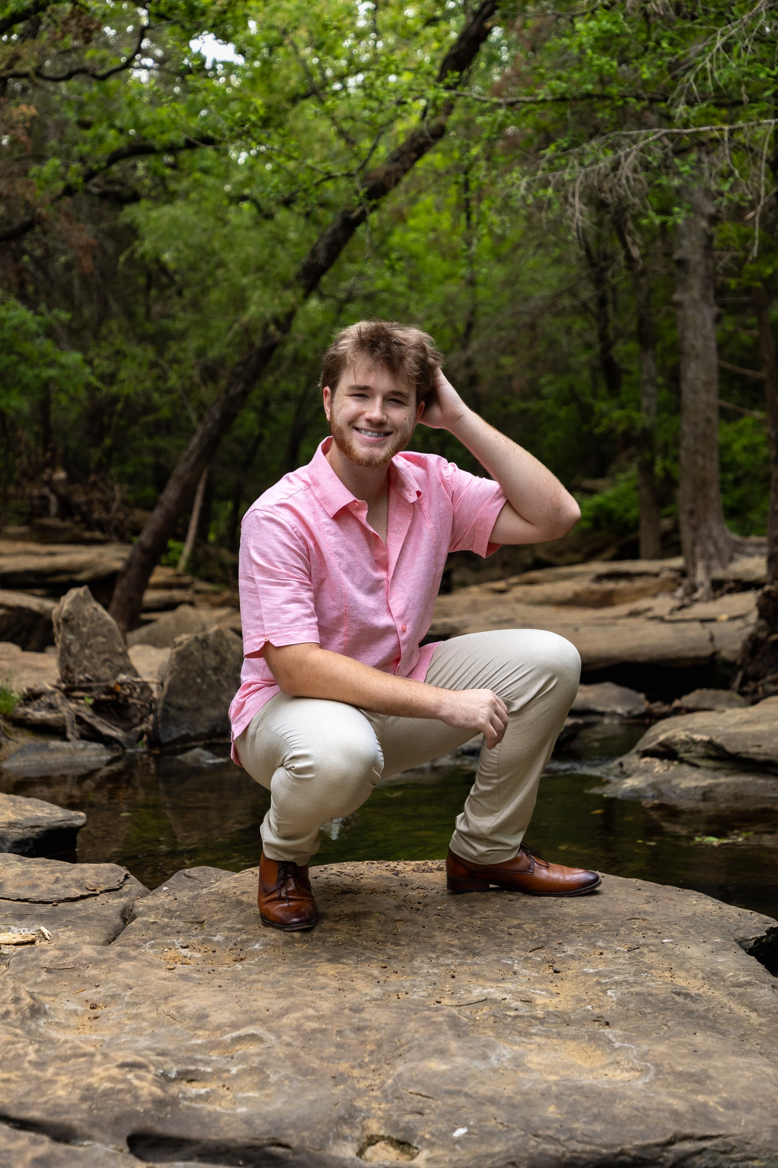 A young man with curly brown hair, a beard, wearing a pink short-sleeve shirt, beige pants, and brown boots, squatting on a large flat rock near a creek in a wooded area with green trees.