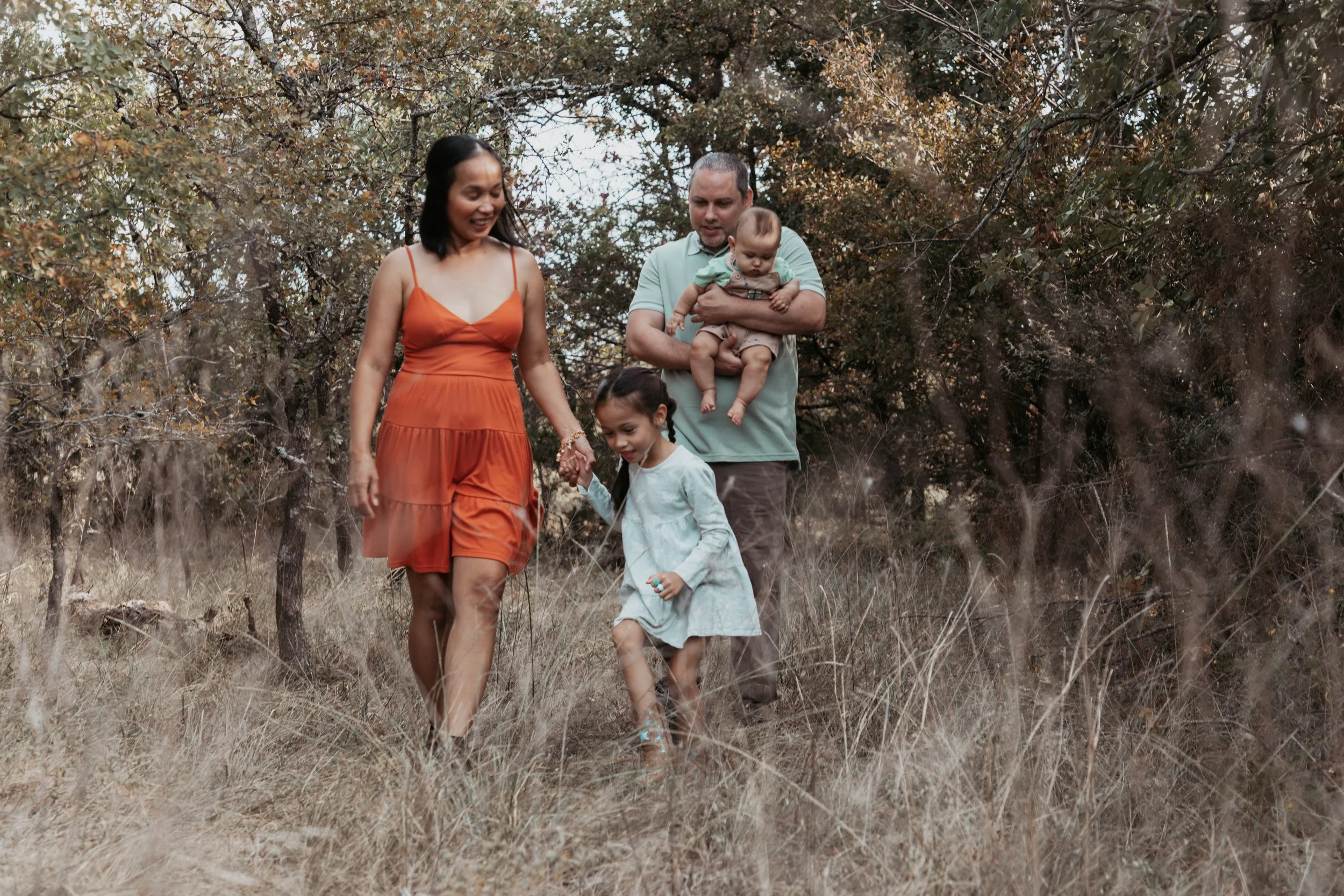 A family of four walking through a grassy, wooded area. The mother in an orange dress, the father holding a baby, and two young girls, one holding the mother's hand, all smiling and enjoying the outdoor walk.