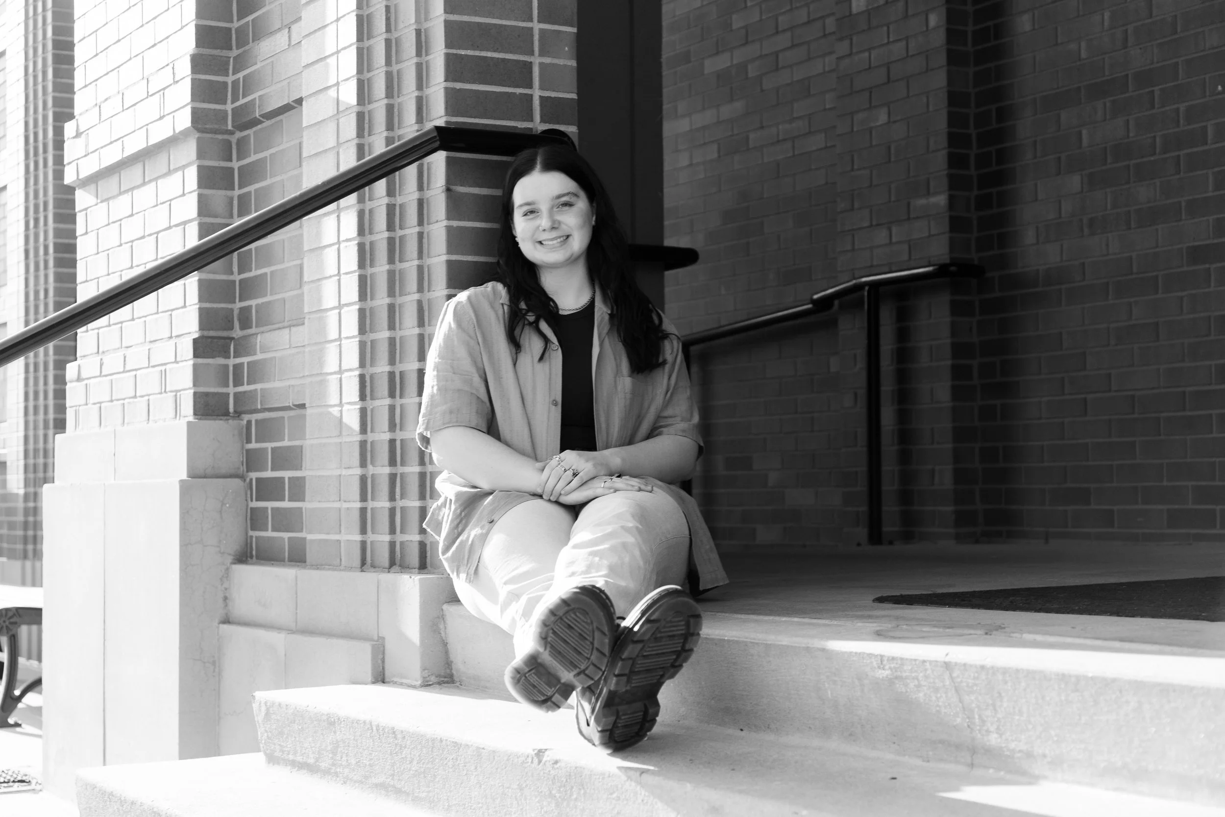 A young woman with dark hair and light skin sitting on stairs outside a brick building, smiling at the camera, wearing casual clothing including a jacket and boots.
